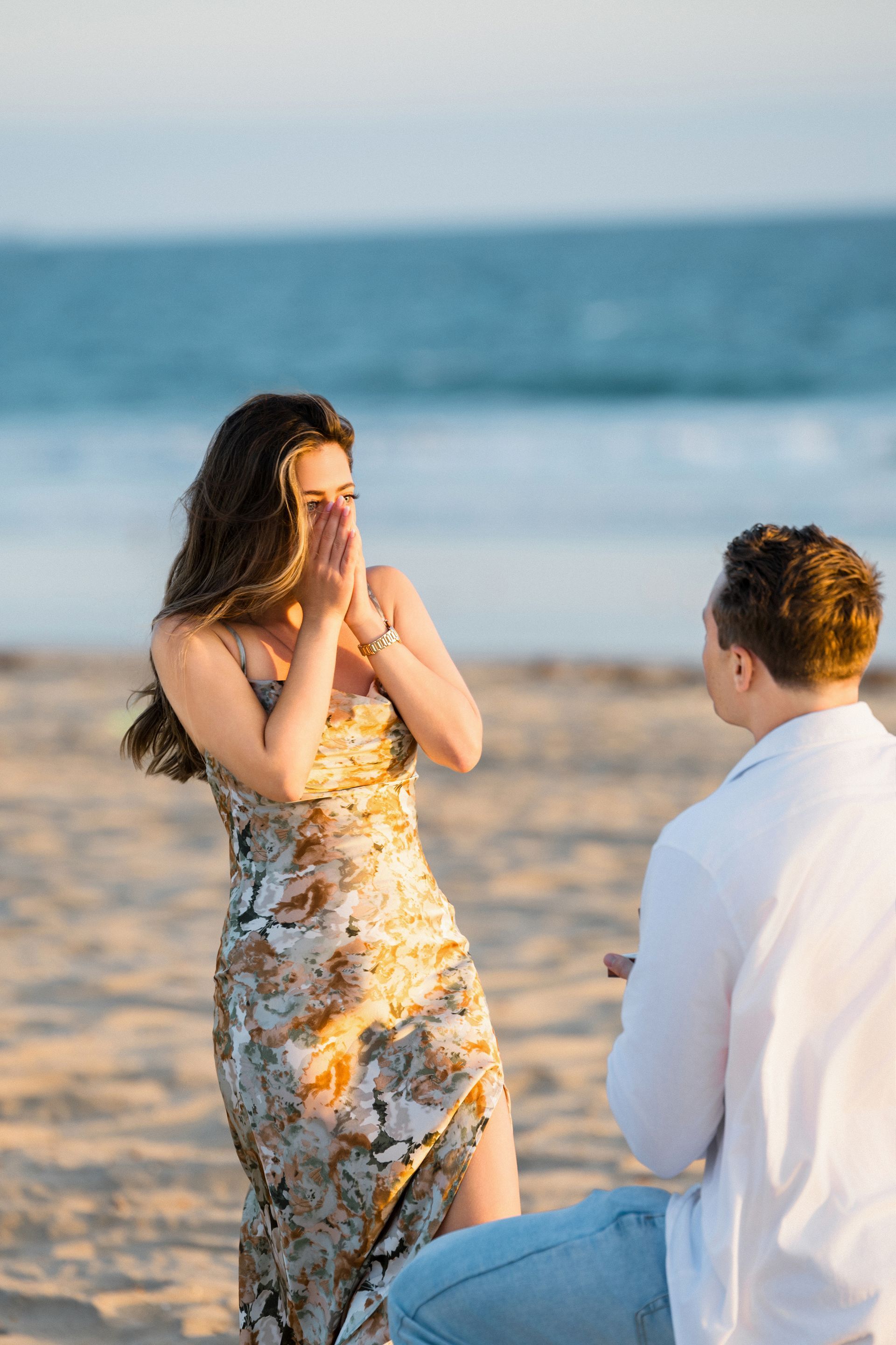 Couple stands on Huntington Beach shoreline during sunset, with the man down on one knee proposing. The woman covers her mouth in surprise, surrounded by warm pink and orange hues reflecting on the ocean waves