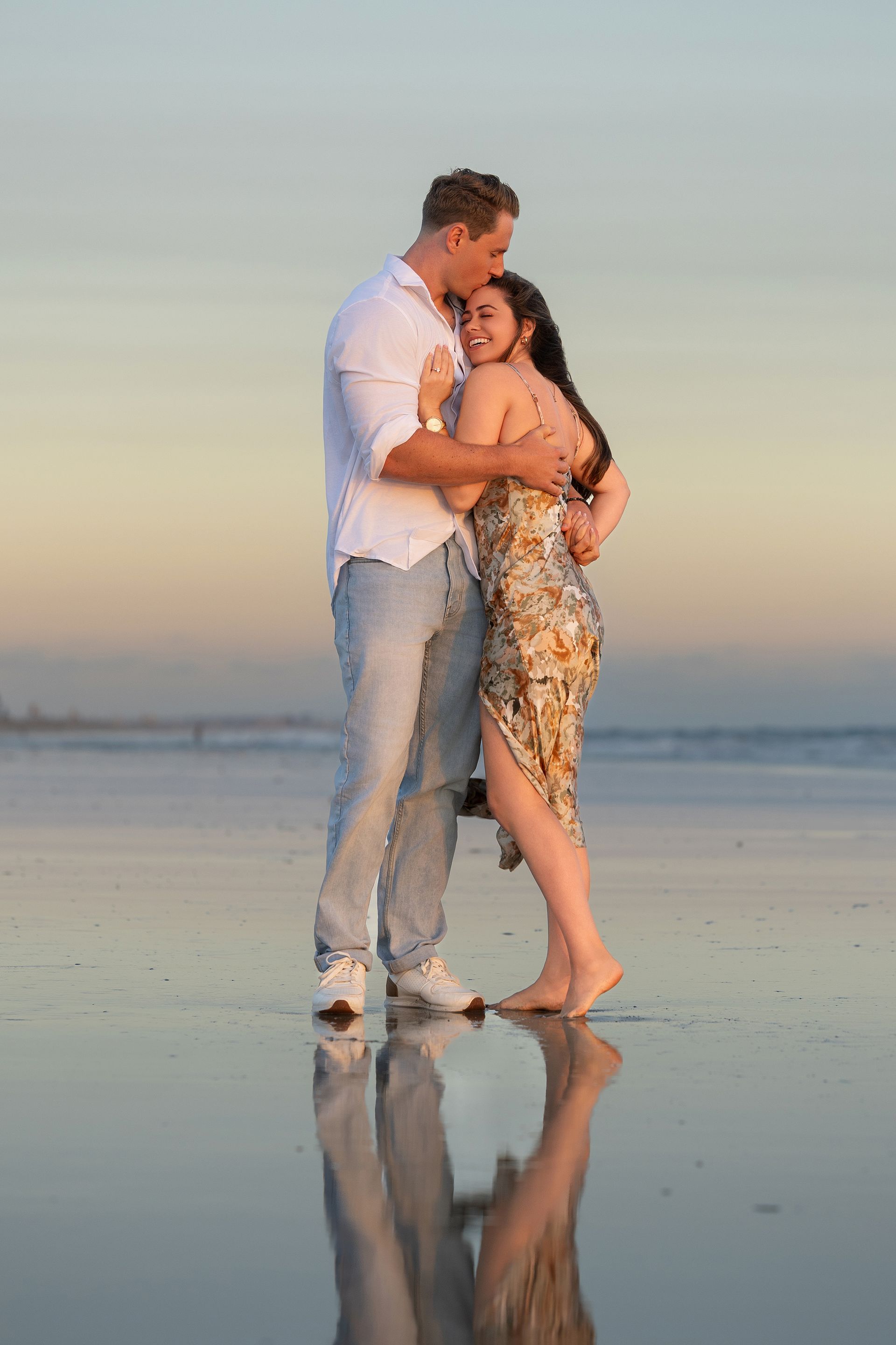 A couple embracing joyfully on Huntington Beach as the sun sets over the ocean, moments after a marriage proposal
