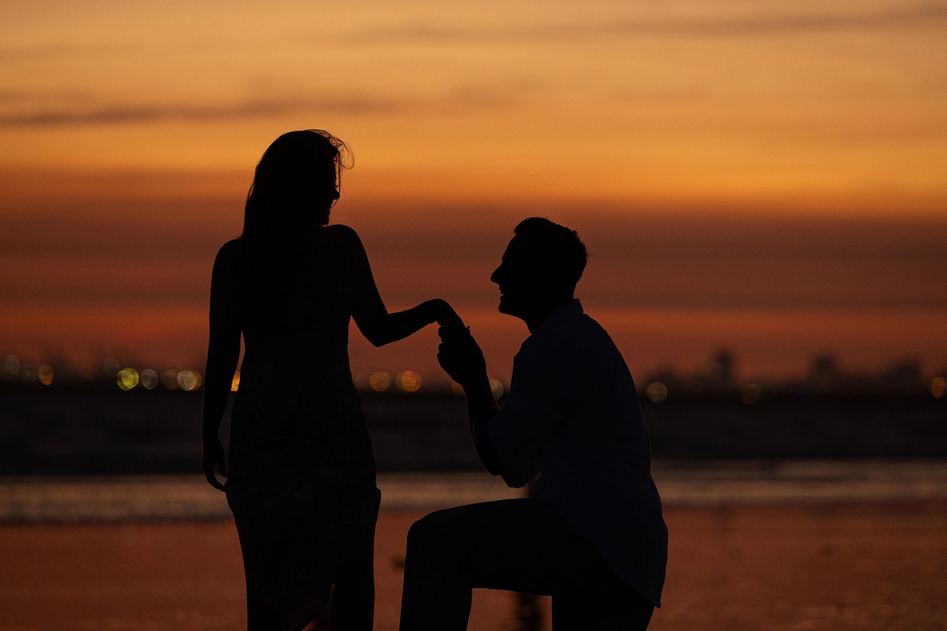 A man on one knee holding an engagement ring while proposing to his partner on the sandy shores of Huntington Beach at sunset
