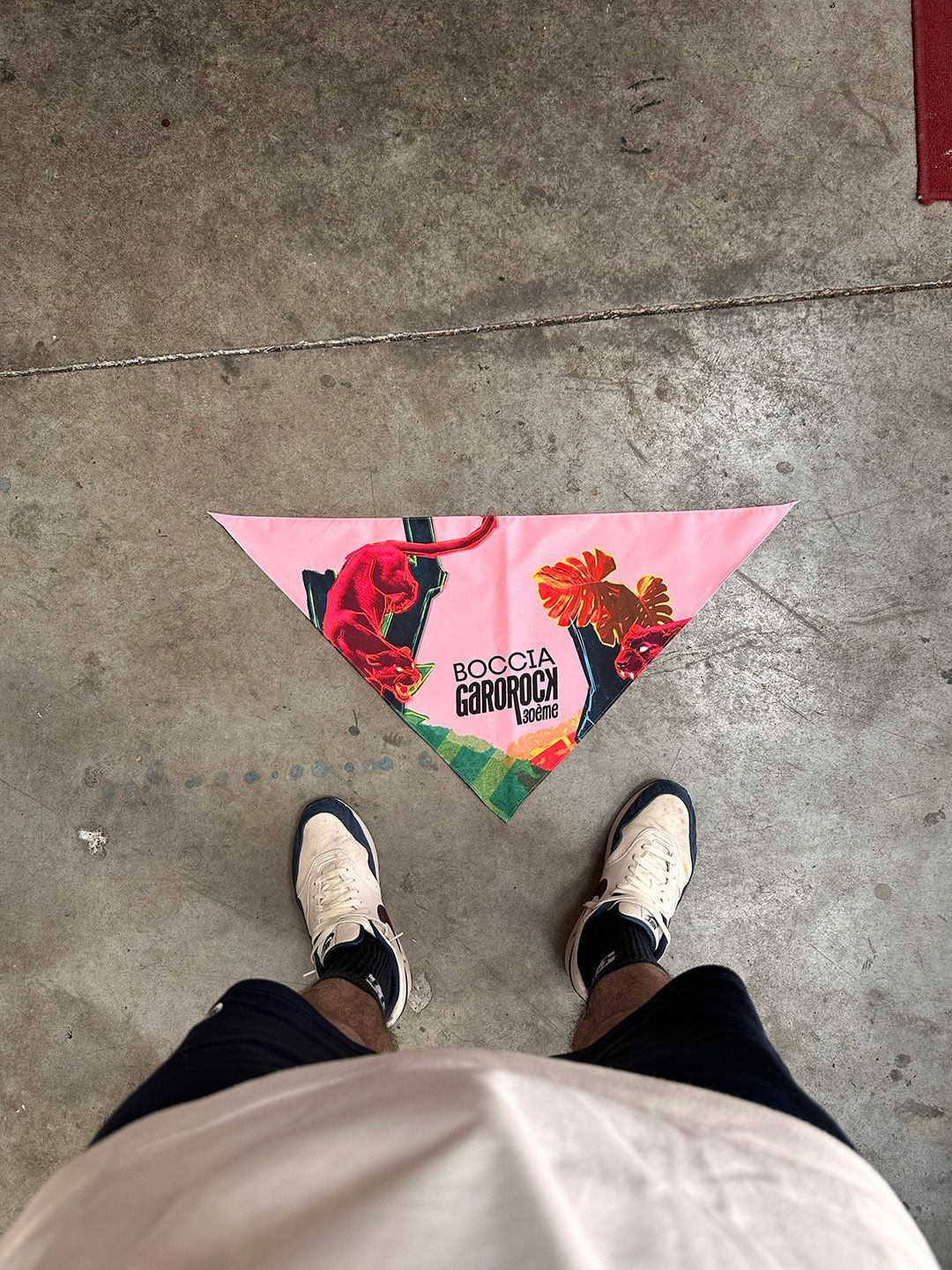 Les pieds d'une personne chaussée de baskets se dressent au-dessus d'une écharpe triangulaire rose à motifs floraux et inscription noire, posée sur le sol.