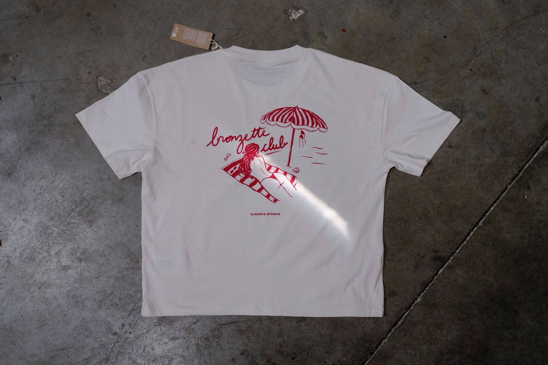 Un t-shirt blanc orné d'une illustration rouge représentant une scène de plage avec une personne sous un parasol et un texte en écriture cursive.