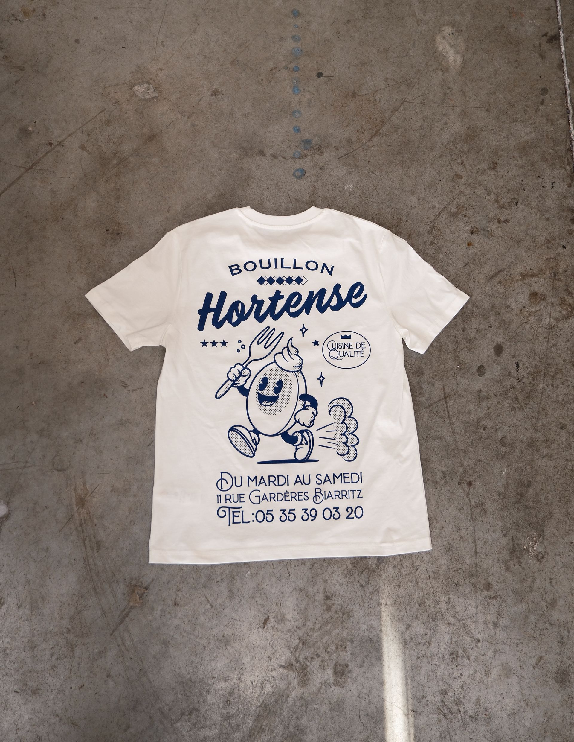 Un t-shirt couleur crème avec un dessin bleu représentant un œuf de dessin animé, le texte « Bouillon Hortense » et un numéro de téléphone.