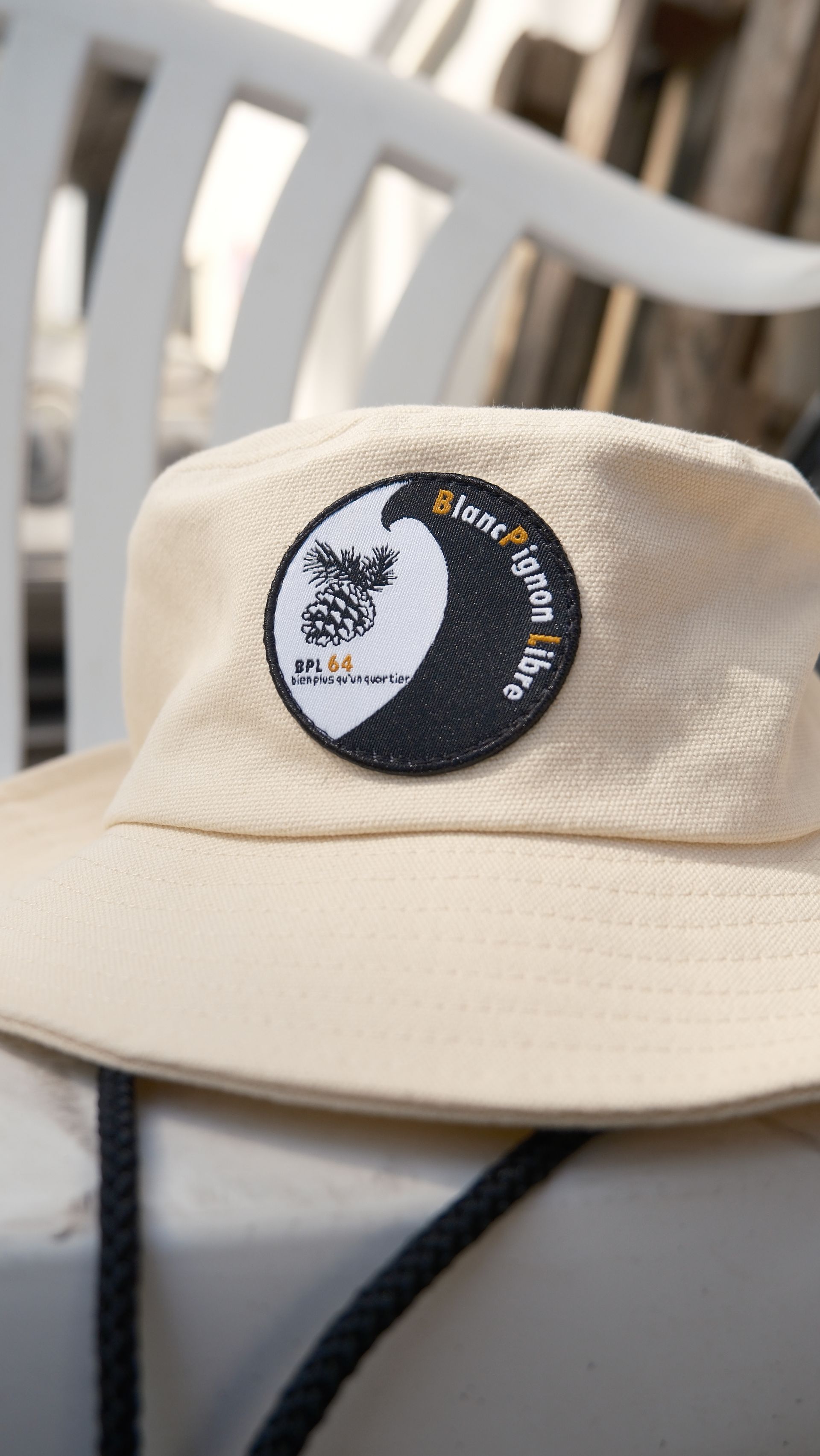 Un bob couleur crème orné d'un écusson circulaire représentant une vague stylisée et du texte, posé sur une chaise blanche.