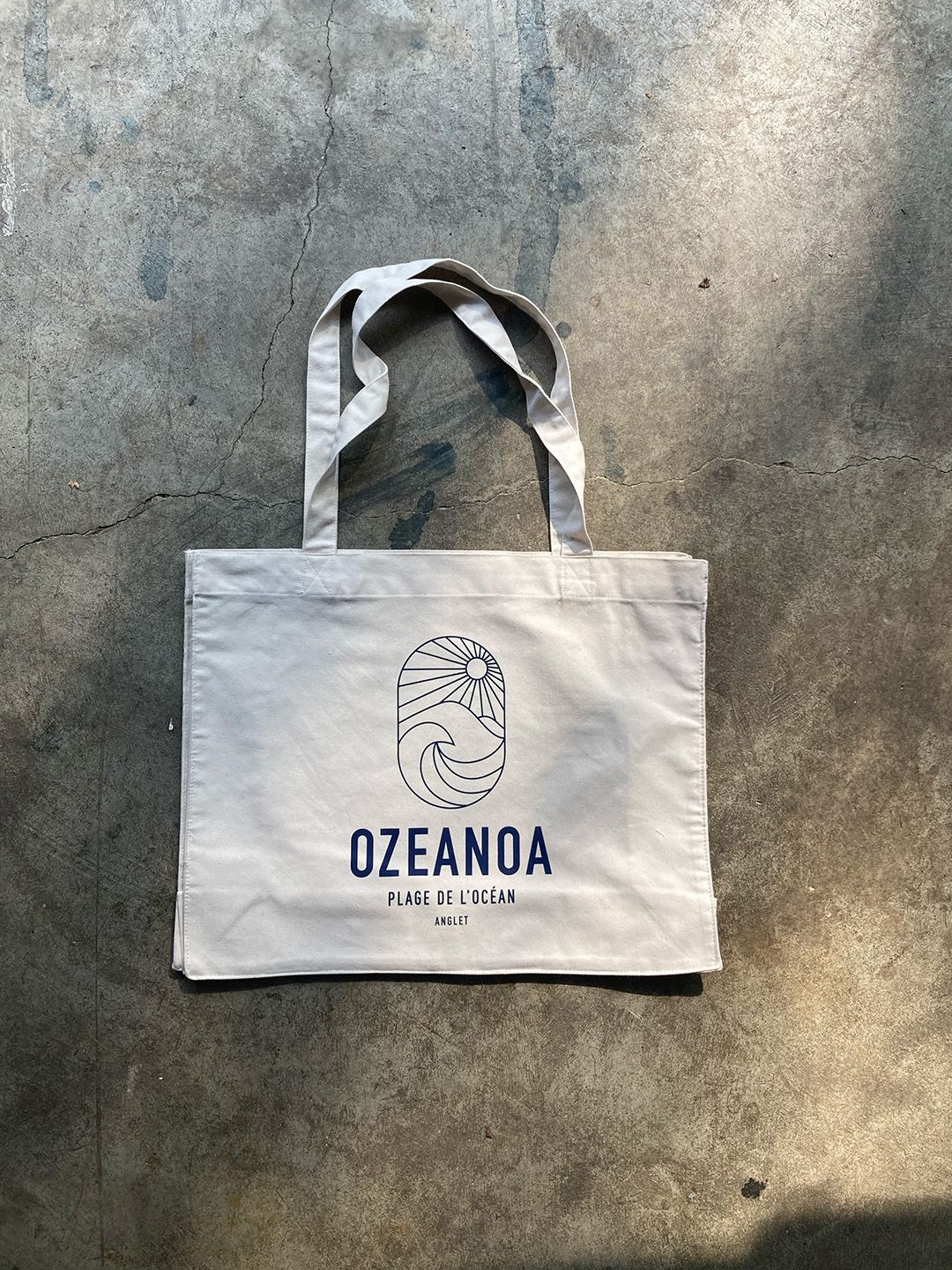 Un sac cabas en toile beige orné d'un logo bleu représentant un soleil et une vague, portant l'inscription « OZEANOA », repose sur une surface en béton gris.