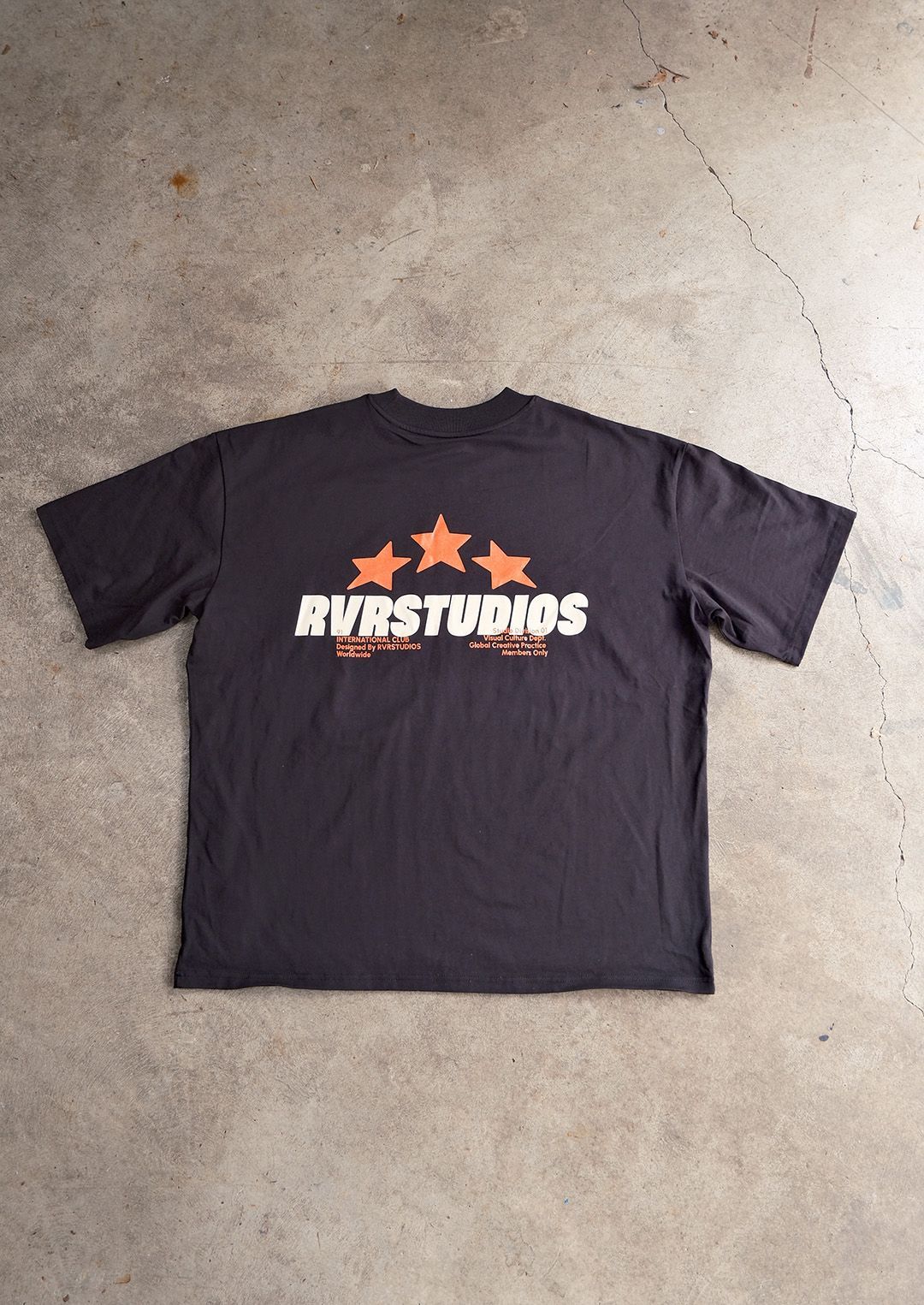 Un t-shirt noir posé à plat sur un sol en béton gris, orné d'un motif à trois étoiles orange au-dessus du texte « AVRSTUDIOS ».