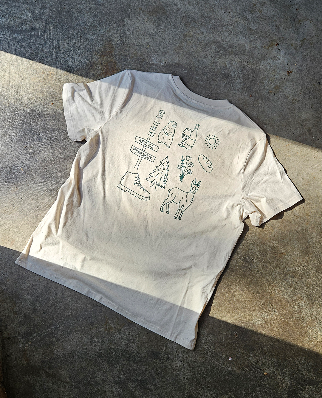 Un t-shirt couleur crème, orné au dos d'une grille de dessins au trait sur le thème de la nature, est posé sur une surface en béton.