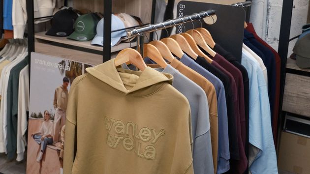 Un portant à vêtements présente une rangée de sweat-shirts à capuche pliés, de couleur beige, grise, bleue et foncée, près d'une affiche publicitaire.