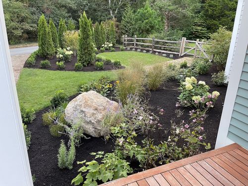 A well-landscaped garden viewed from a porch. Features include a large boulder, green grass, dark mulch, and a small wooden fence.