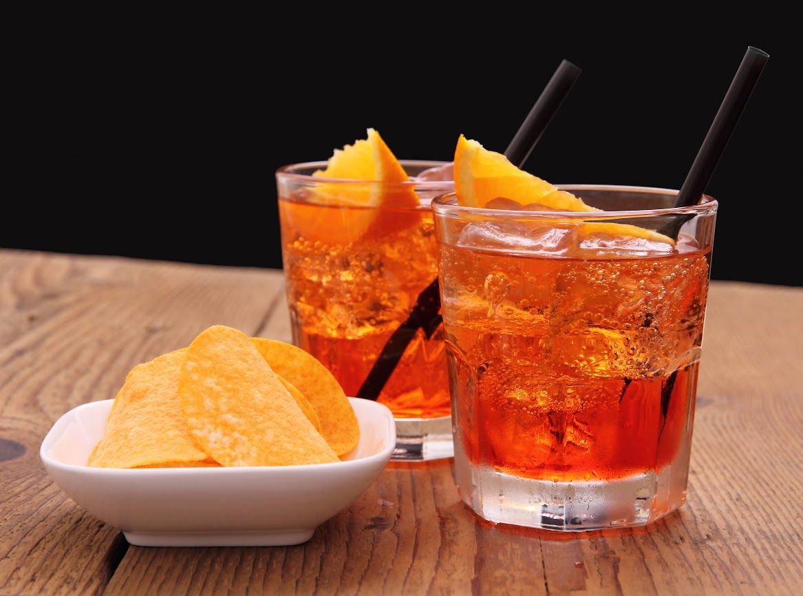 Due bicchieri di Aperol Spritz all'arancia con fette di agrumi e una ciotolina di patatine fritte su un tavolo di legno.