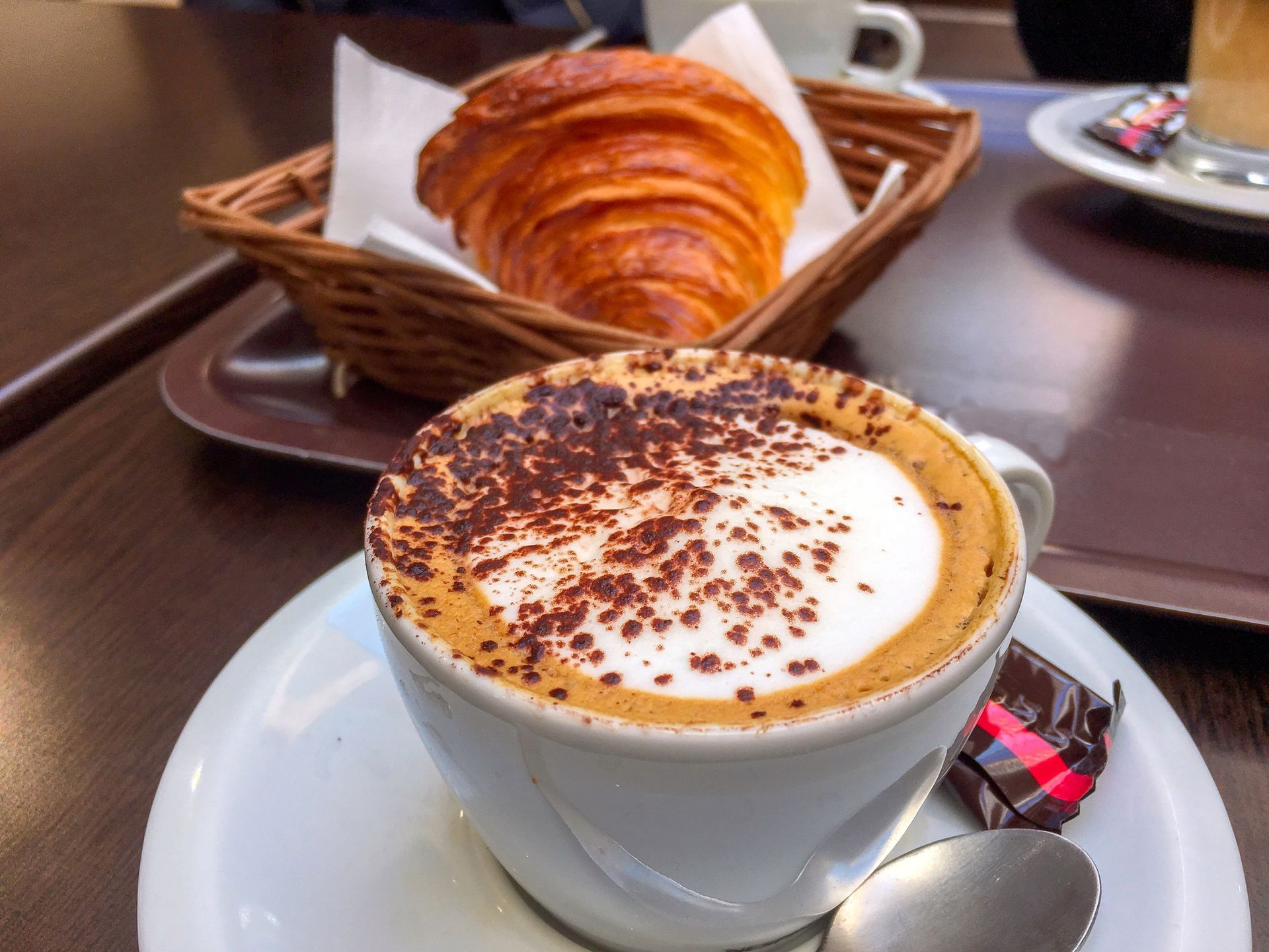 Una tazza di cappuccino con una spolverata di cacao, adagiata accanto a un croissant in un piccolo cestino di vimini su un vassoio marrone.