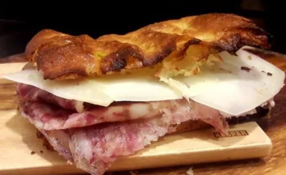 Un panino gourmet con salumi e formaggio tagliato finemente su pane piatto tostato, adagiato su un tagliere di legno.