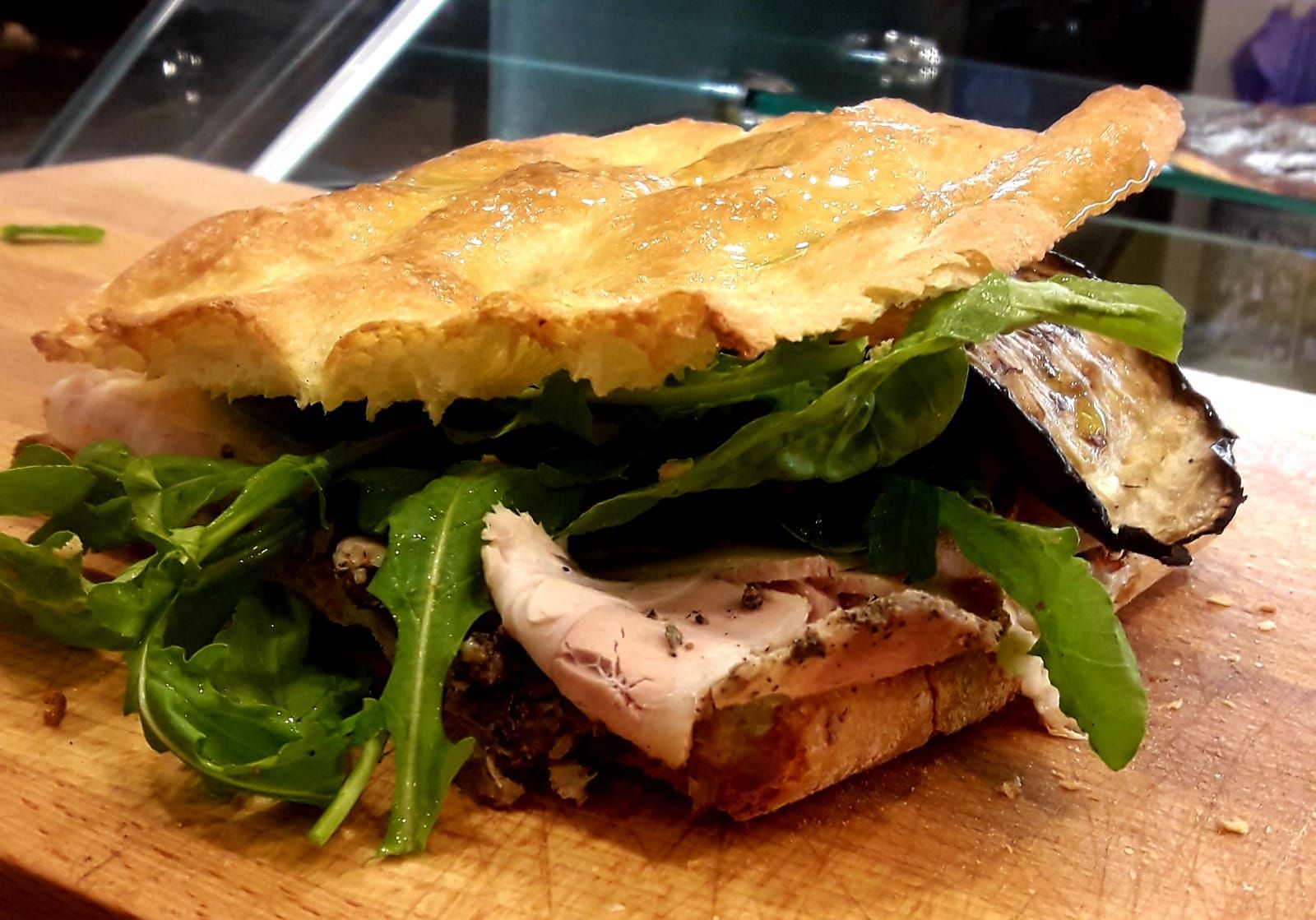 Un panino di focaccia su un tagliere di legno, farcito con rucola, affettati e una fetta di melanzana grigliata.