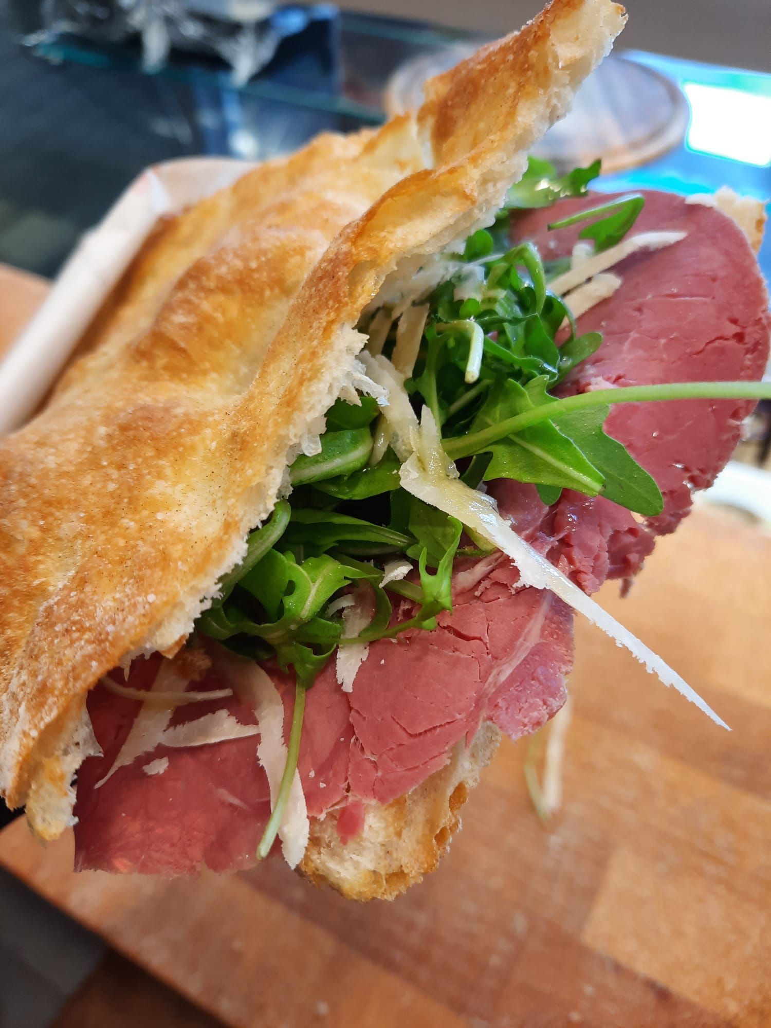 Un panino con roast beef su pane ciabatta con rucola fresca e scaglie di parmigiano, servito su un tagliere di legno.