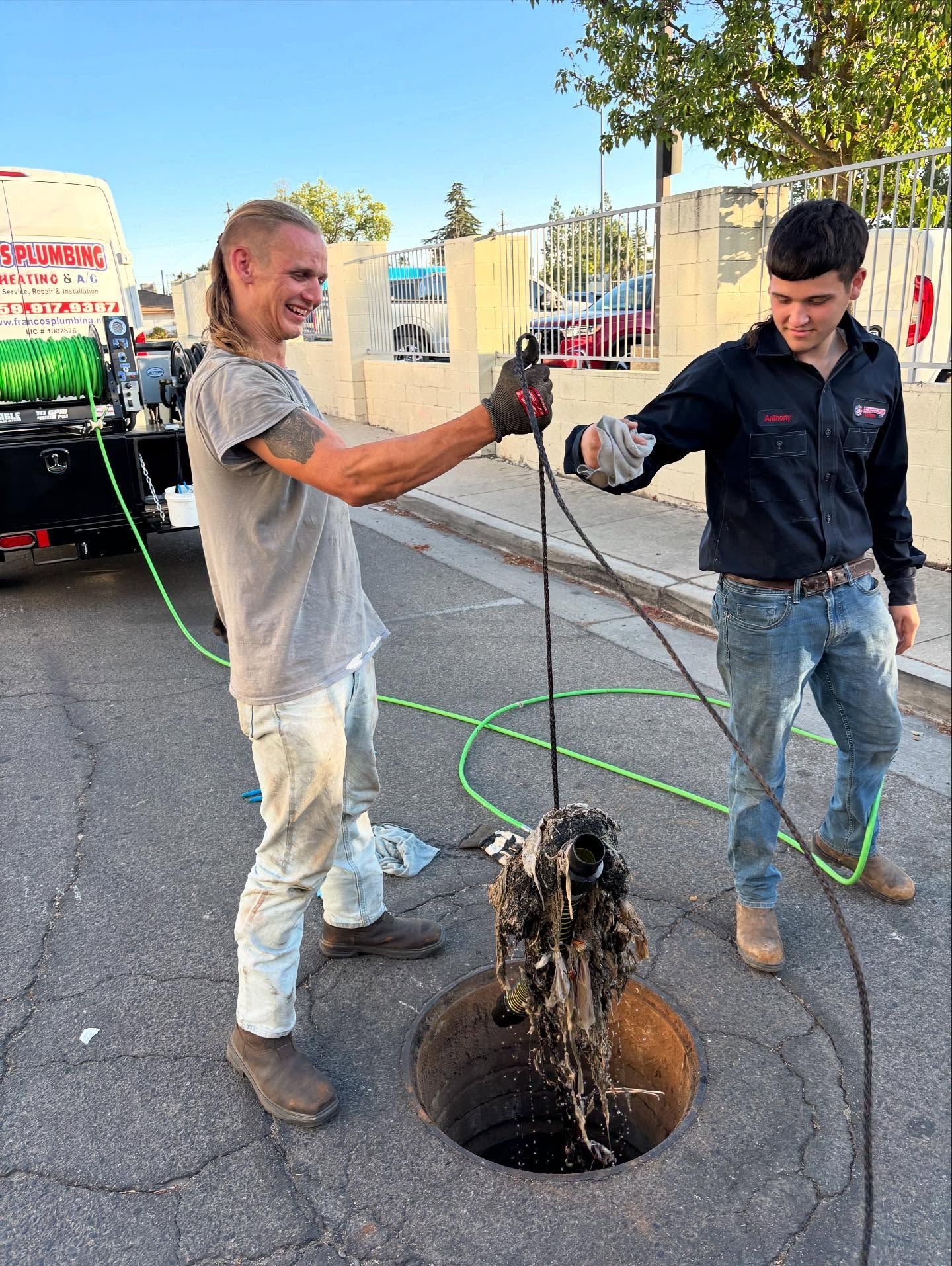 Two men clearing a clogged sewer. One pulls a dirty object from the manhole, smiling; the other holds the hose.