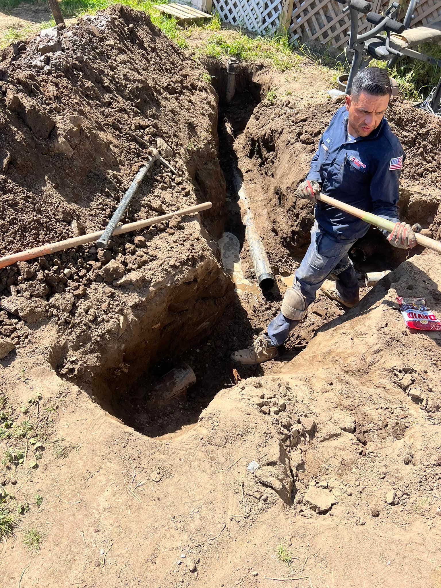 Man in uniform repairs pipe in trench, holding a shovel, outdoor setting.