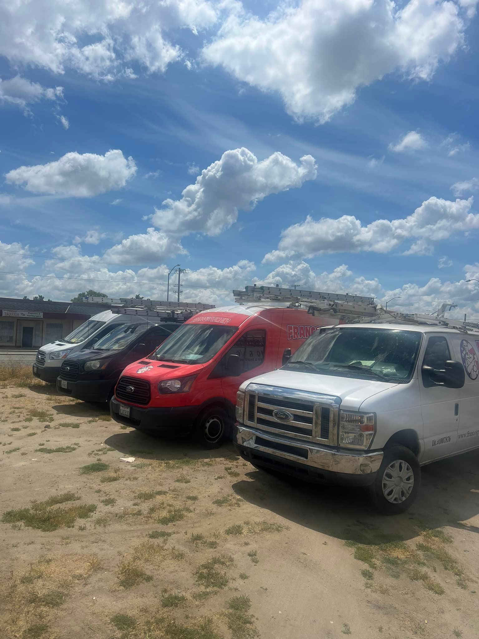 Fleet of service vehicles parked outdoors under a bright blue sky with scattered clouds.