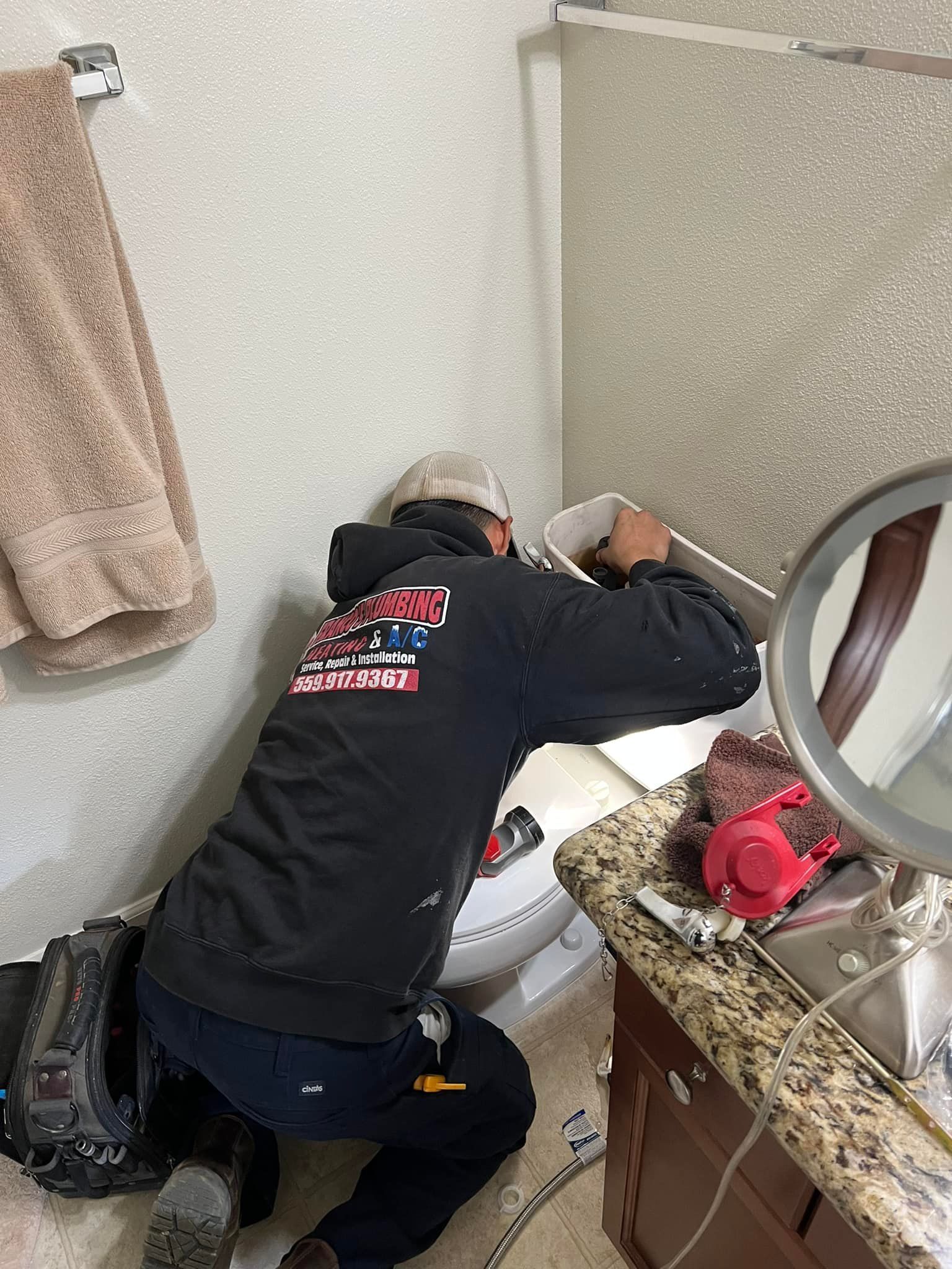A plumber in a black hoodie kneels, working on a toilet. Bathroom setting, speckled wall.
