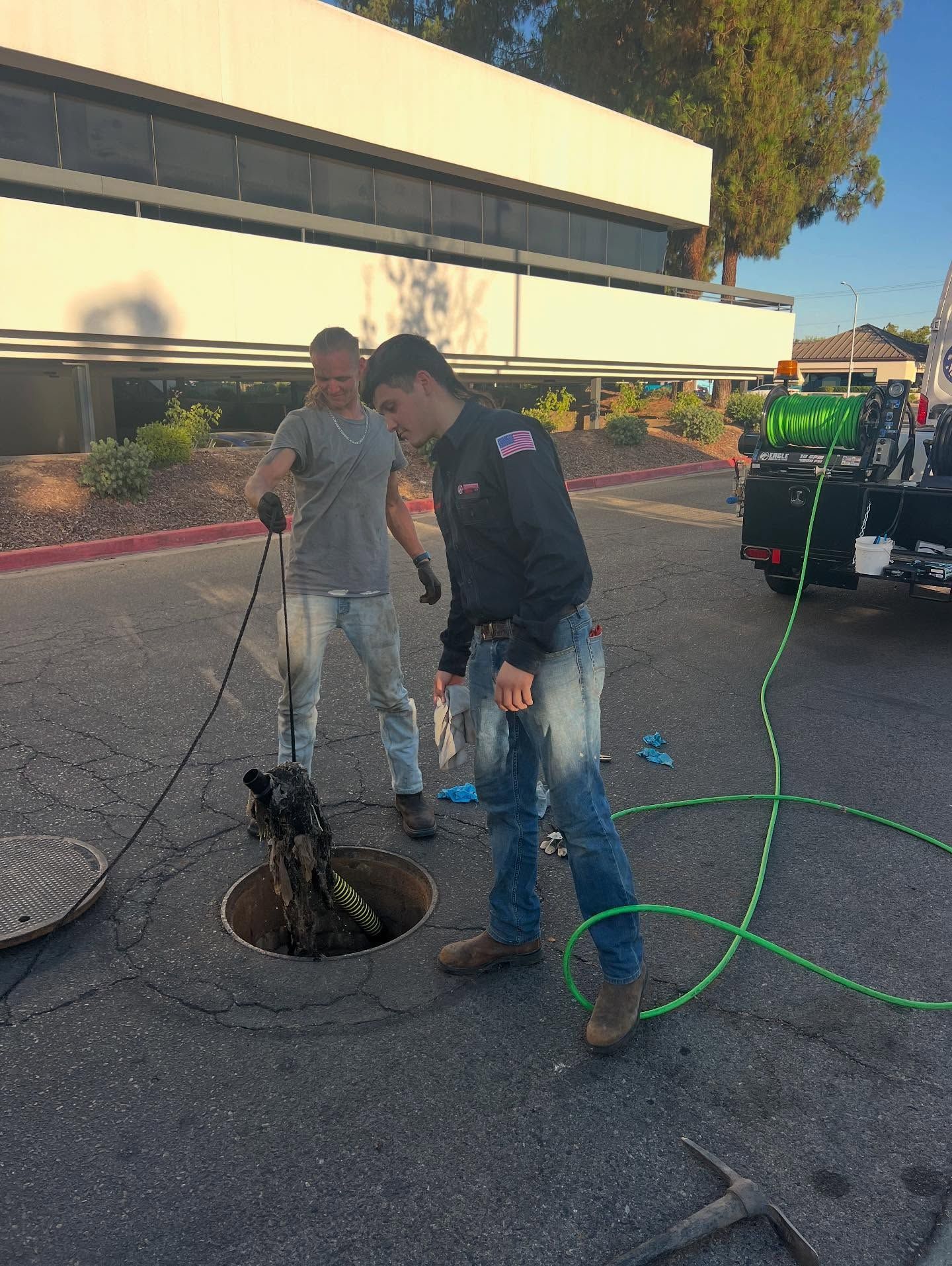 Two men inspecting a sewer manhole in a parking lot; one holds a cable, the other looks inside.