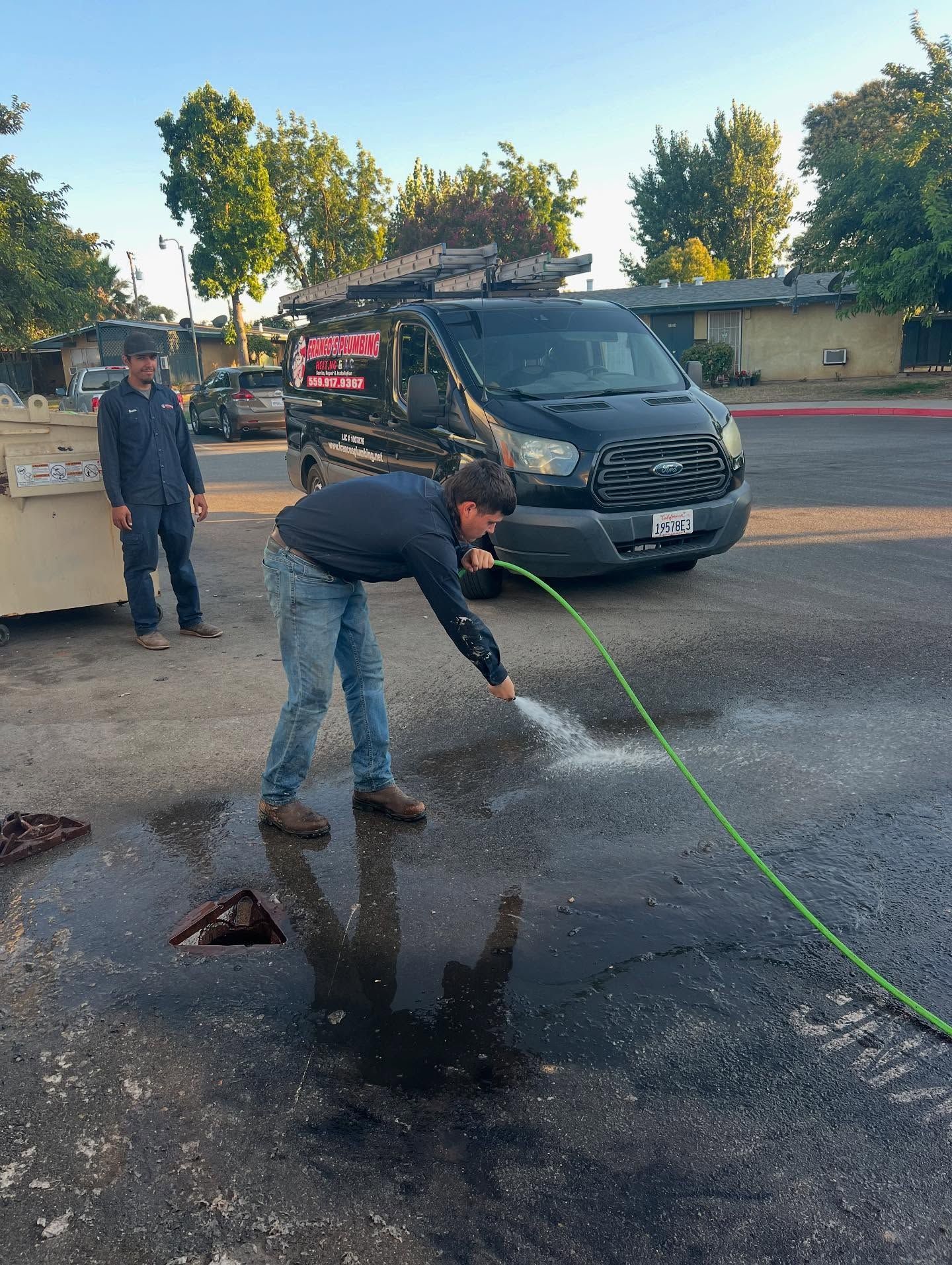 Man sprays water with a hose on a dark, oily patch in a parking lot; another man watches near a van.