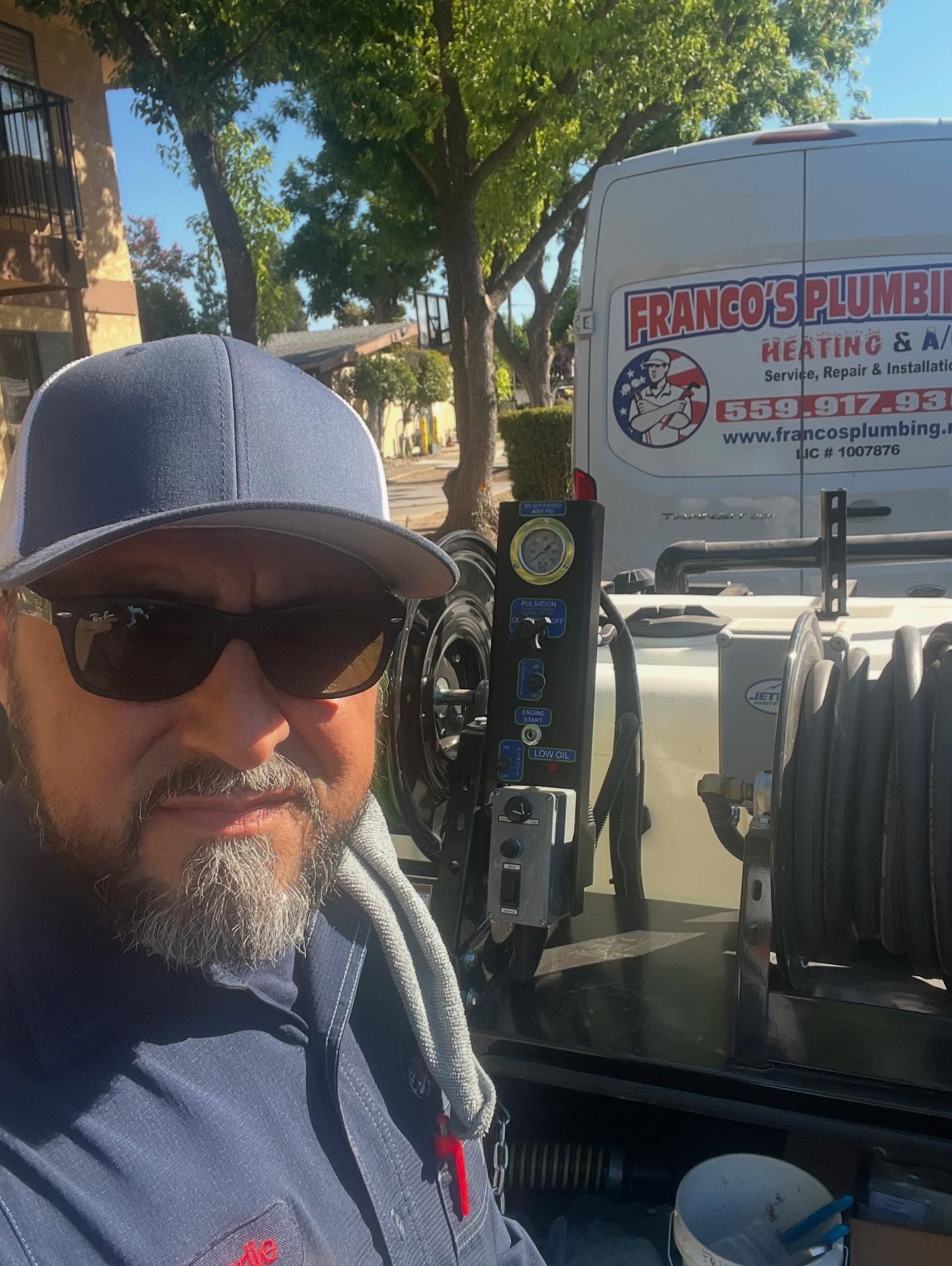 Man in sunglasses and cap stands in front of a white plumbing truck, sunny outdoors.