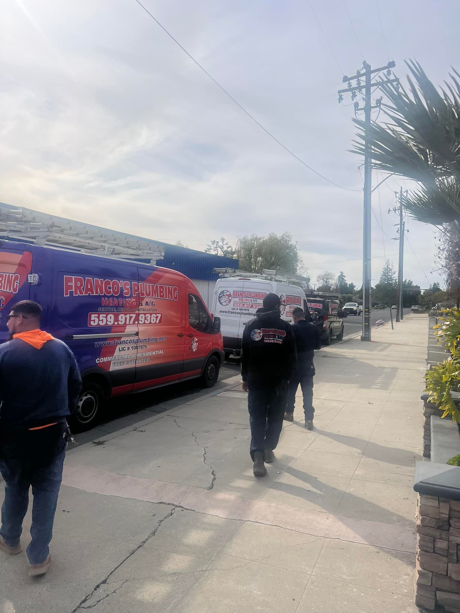 People walking alongside service vans on a sidewalk in front of a house; sunny day.