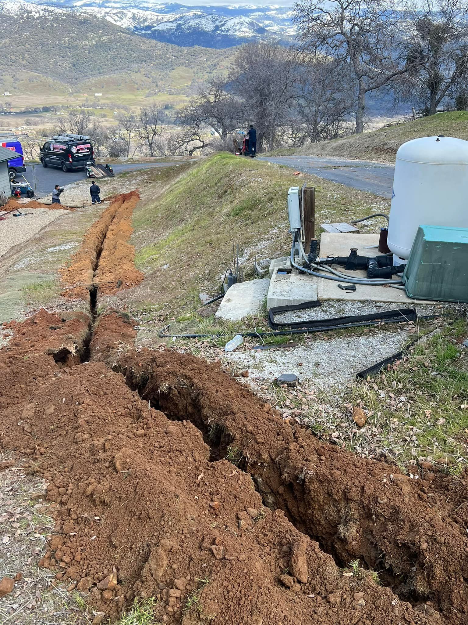 Trenches dug near a water system with a tank and machinery, hillside setting with vehicles and people.