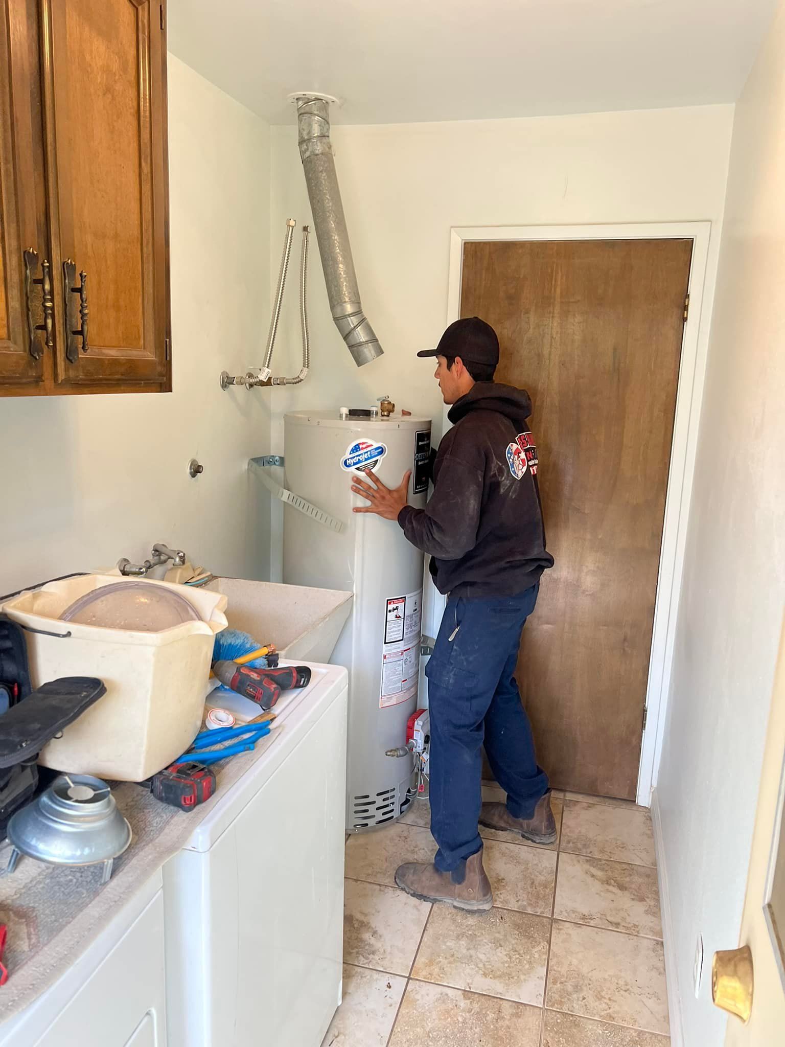 A person installs a water heater in a laundry room with tools and supplies on the counter.