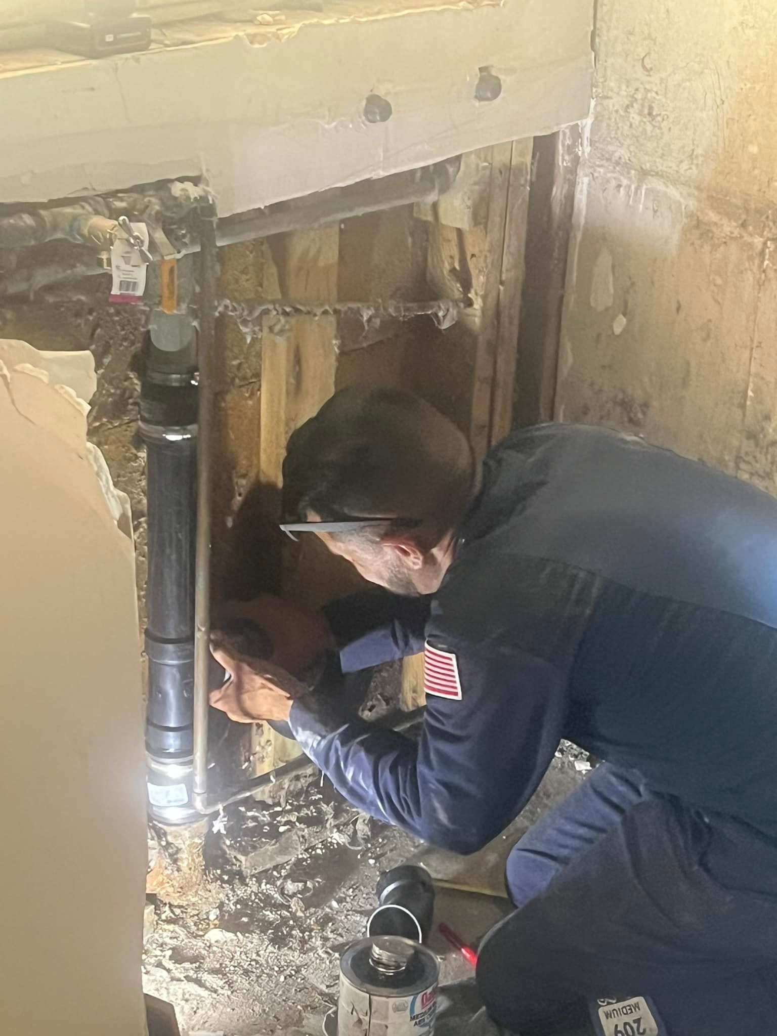 Plumber repairs pipes in a wall. He wears a blue shirt, hat. Exposed wood, pipes visible.