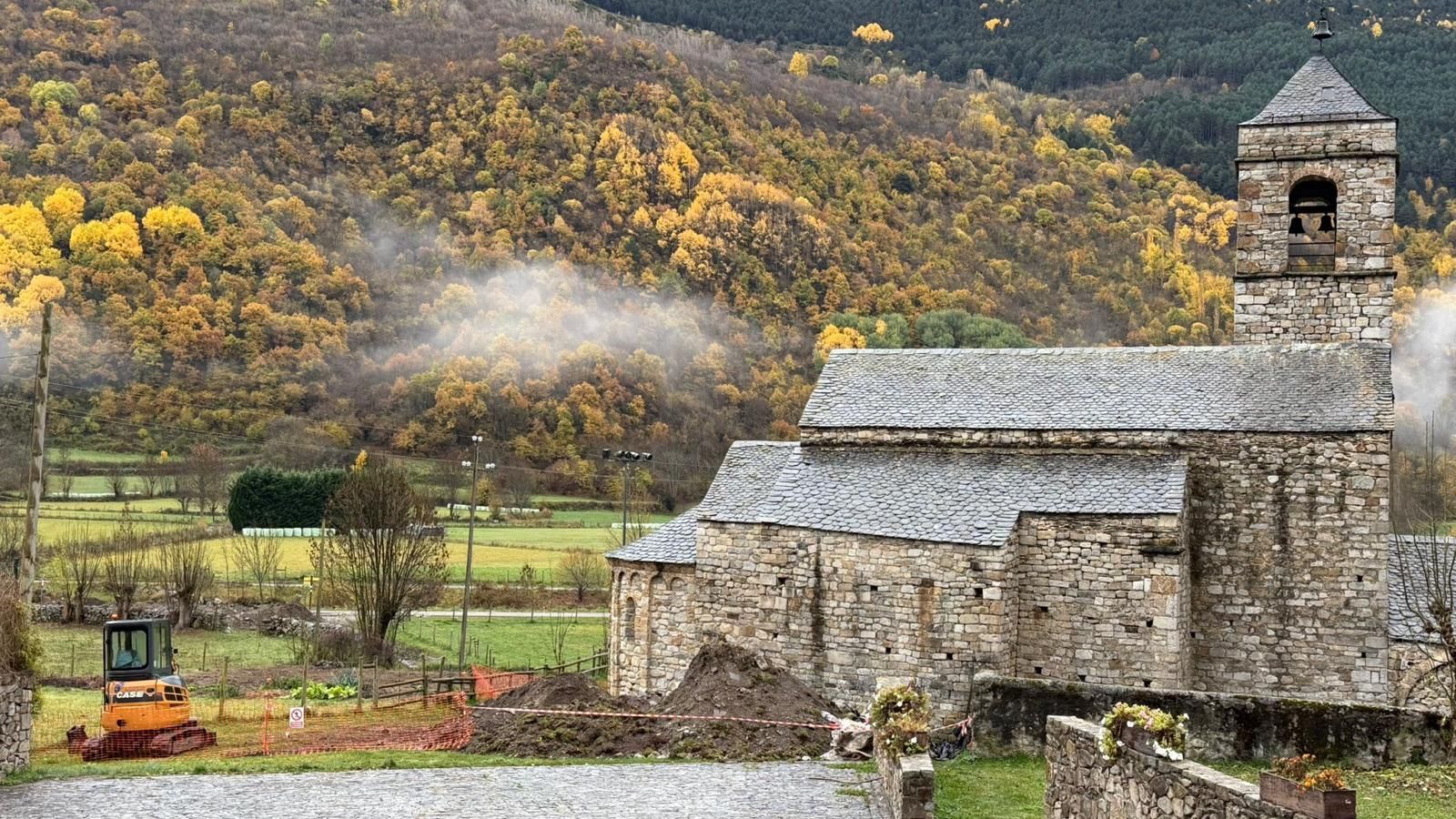 Església de pedra amb campanar en una vall amb fullatge de tardor, un petit vehicle de construcció en primer pla.