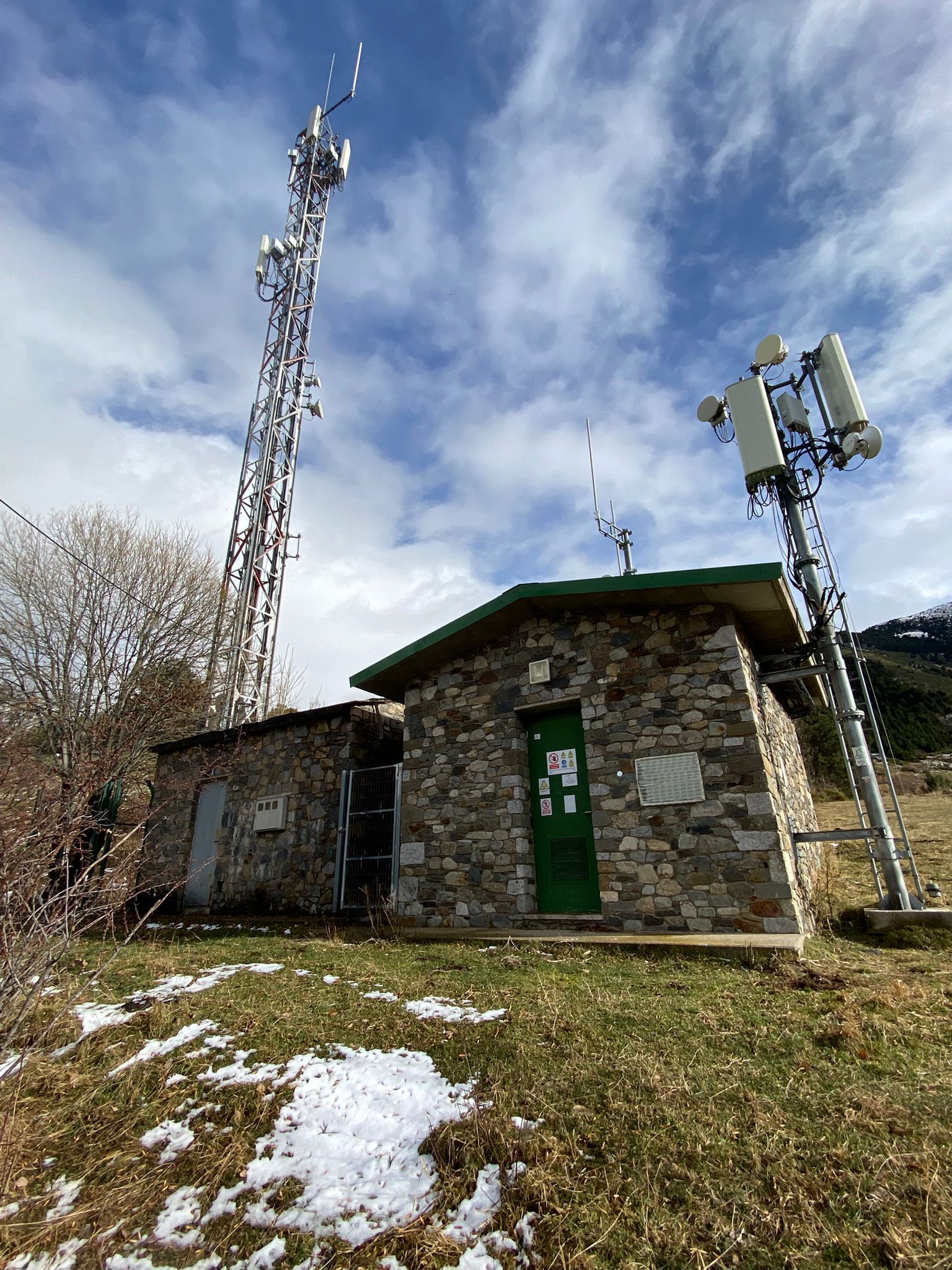 Edifici de pedra amb porta verda i torres d'antena contra el cel ennuvolat.