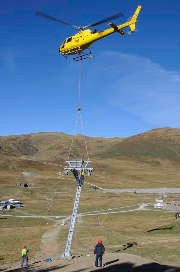 Helicòpter groc aixecant una torre de remuntador d'esquís a una muntanya. Treballadors amb equip de seguretat observen a sota.