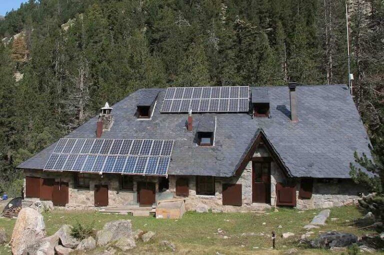 Cabana de pedra amb panells solars sobre una teulada de pissarra, situada en un paisatge de muntanya amb arbres.