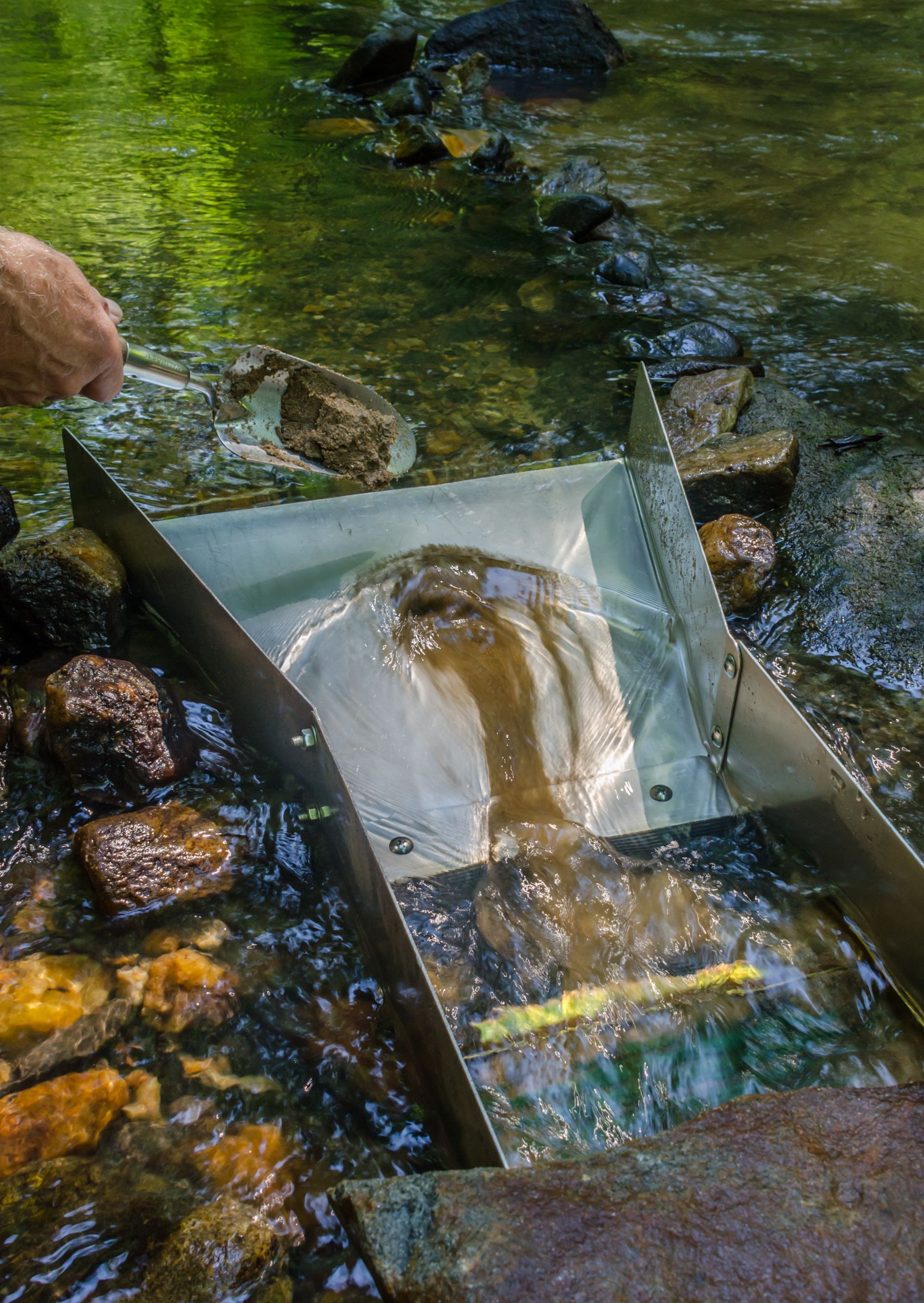 A person is using a metal box to scoop water out of a stream.