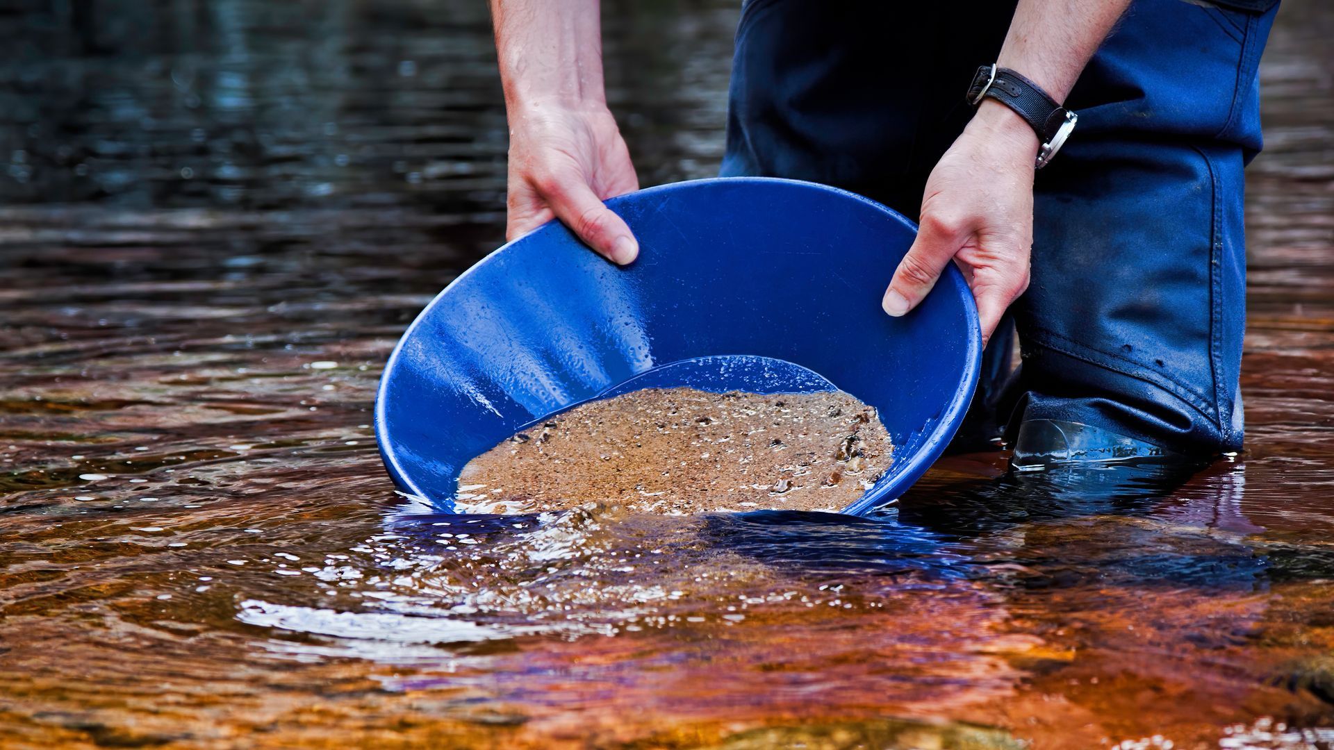 A person is kneeling in the water holding a blue bowl filled with dirt.