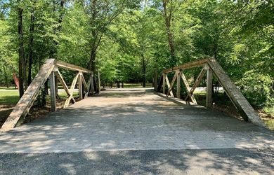 A wooden bridge over a dirt road in a park surrounded by trees.