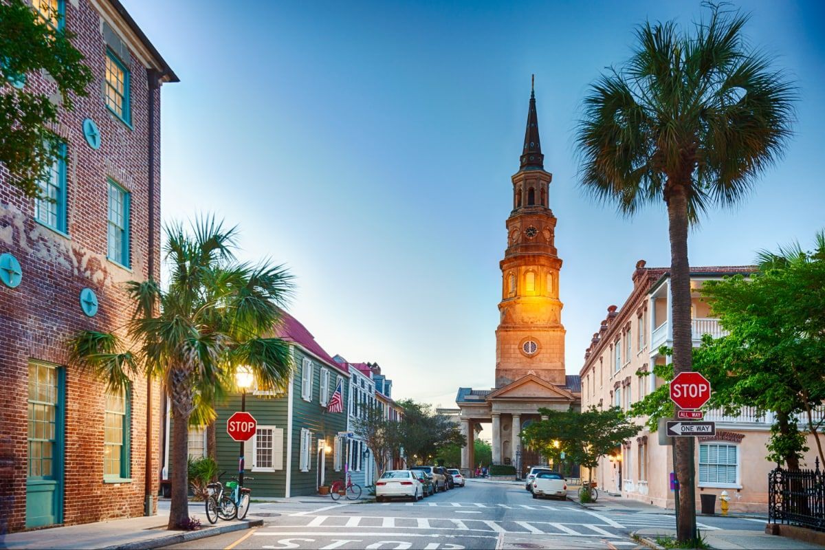 Charleston street with church steeple, palm trees, colorful buildings, and parked cars under a blue sky.