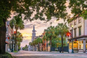 Street in Charleston, SC, with palm trees, historic buildings, and a church spire at sunset.
