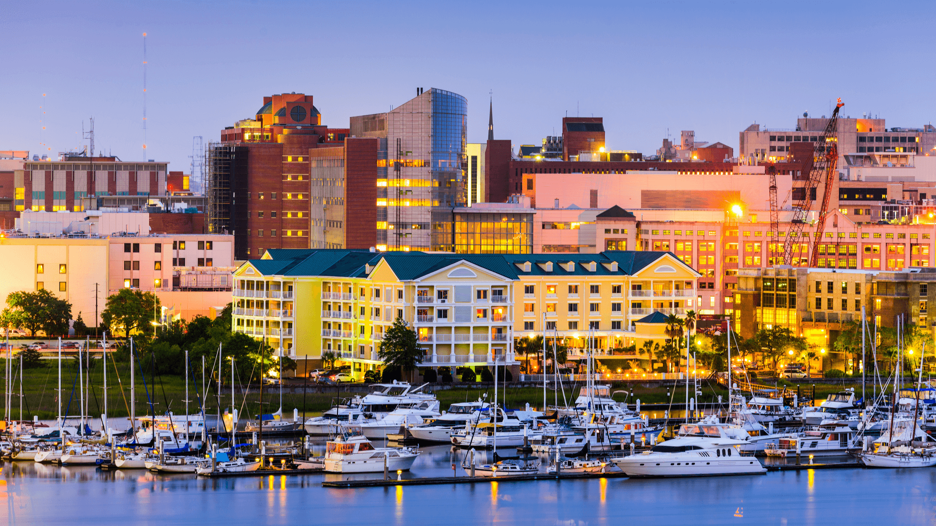 City skyline at dusk with boats docked in harbor, featuring buildings with illuminated lights.