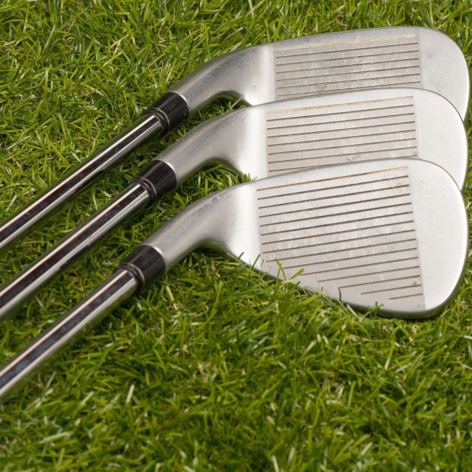 Three silver golf clubs with metal clubheads and shafts lying on a green grass lawn.