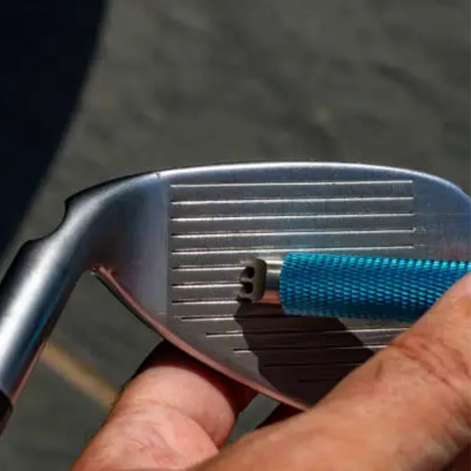 A hand uses a blue groove sharpening tool to clean or restore the metal grooves on a golf club face.
