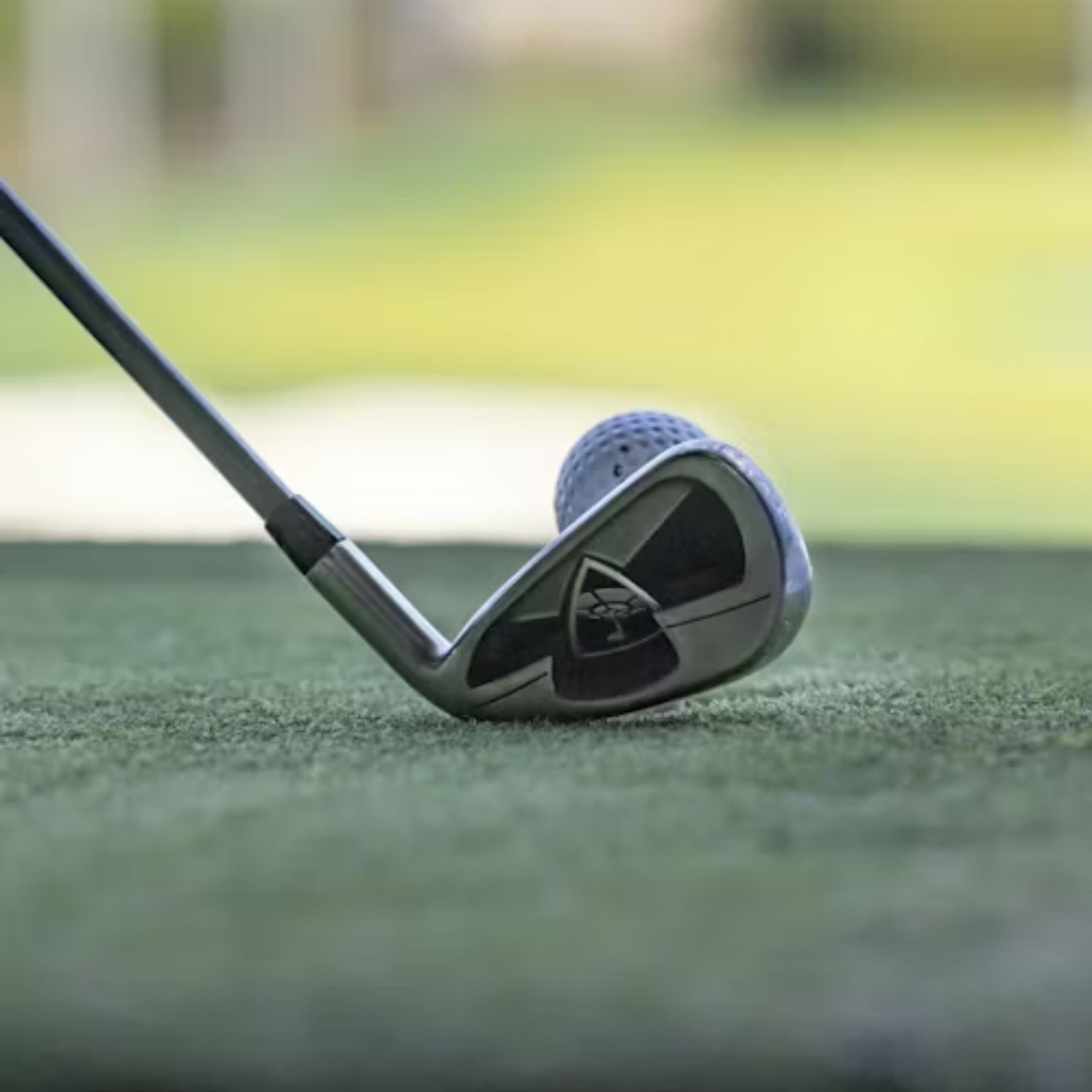 A close-up of a metal golf club head positioned directly behind a white golf ball on a green turf practice mat.