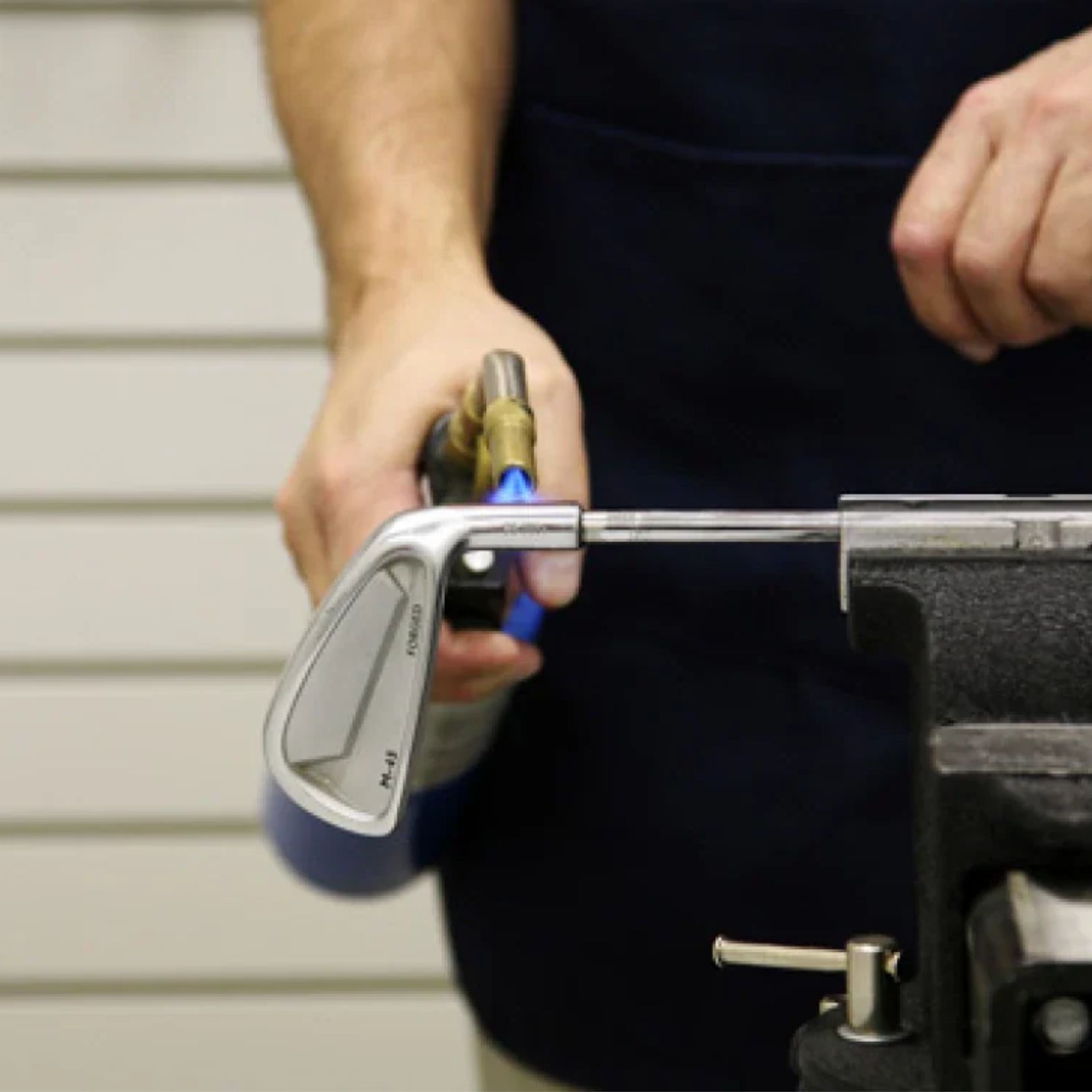 A hand uses a blowtorch to heat the hosel of a golf club held in a metal vise to loosen the epoxy for shaft removal.