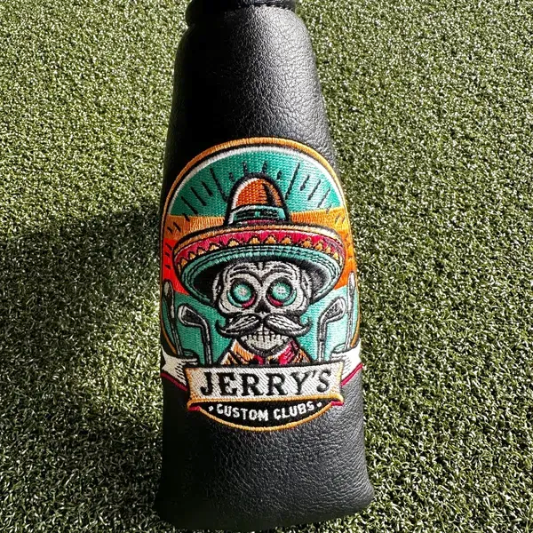 A black golf club headcover features an embroidered logo of a sugar skull wearing a sombrero over a golf club design.
