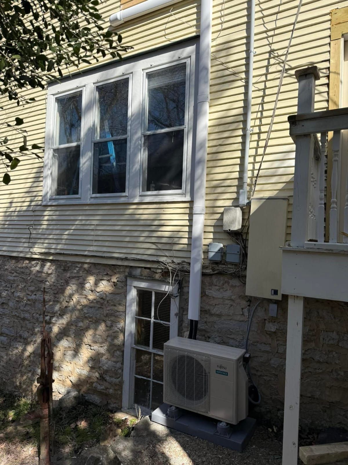 Exterior wall of a house with windows, air conditioner, and white gutter. Stone foundation and yellow siding.