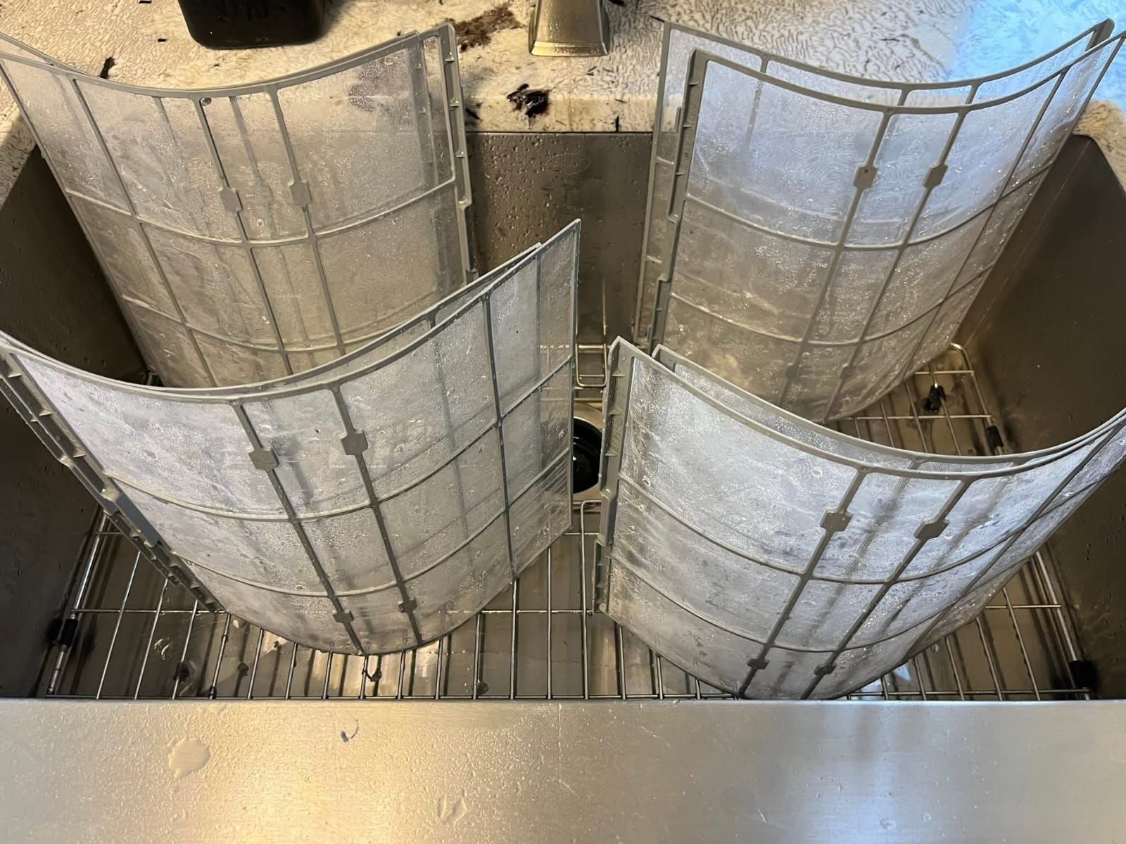 Four dirty air conditioner filters soaking in a stainless steel sink.