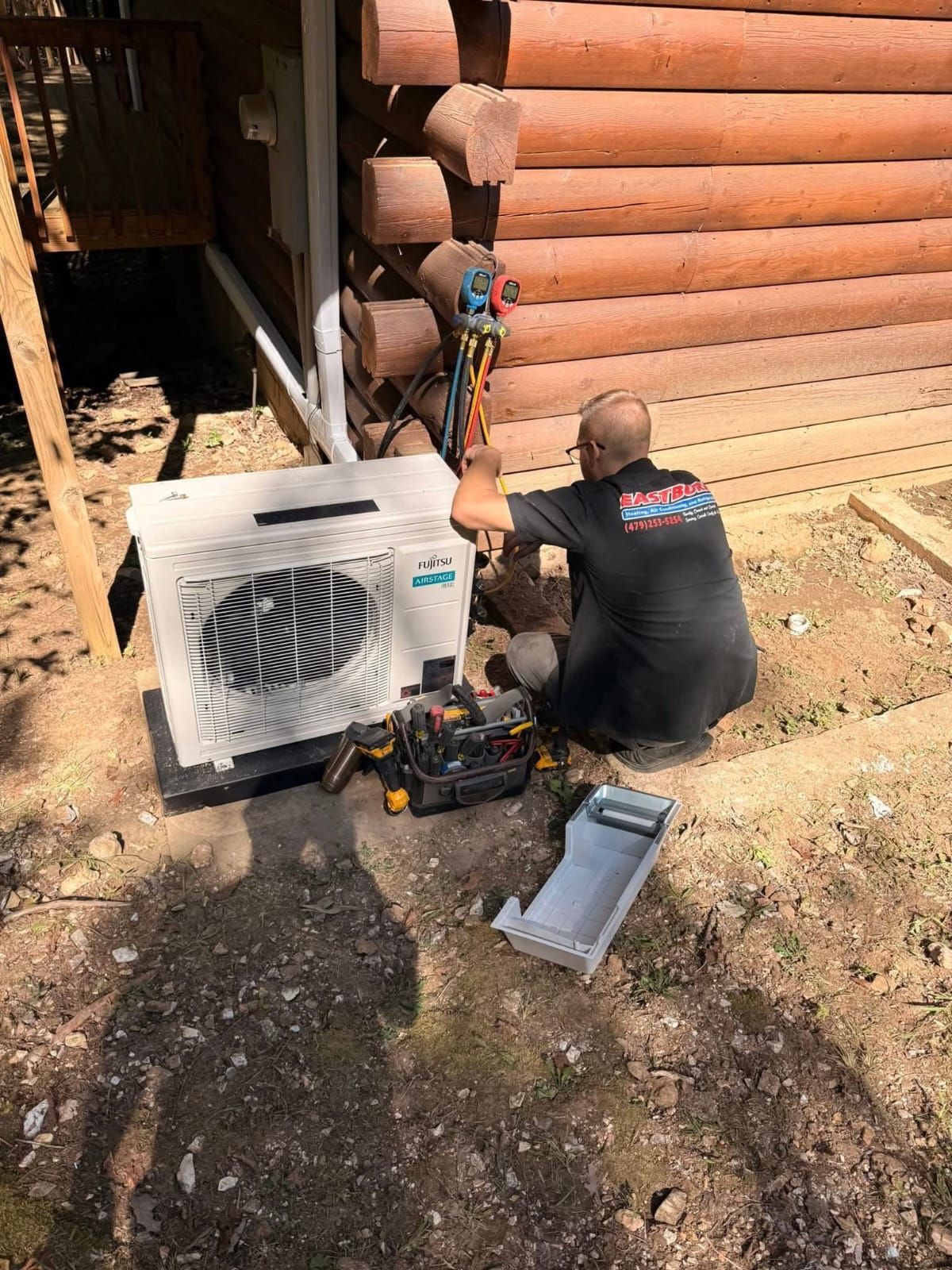 HVAC technician works on an air conditioning unit outside a log cabin. Tools on the ground.