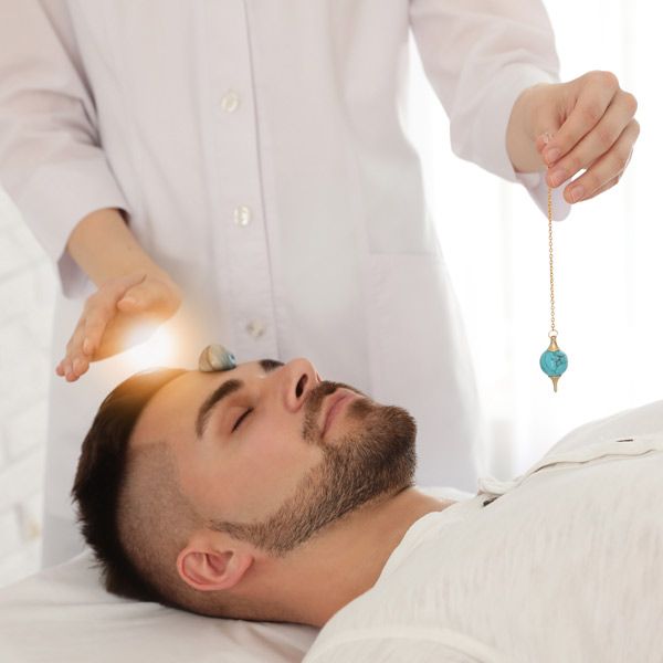 A man is lying on a bed while a woman holds a crystal pendulum over his head.