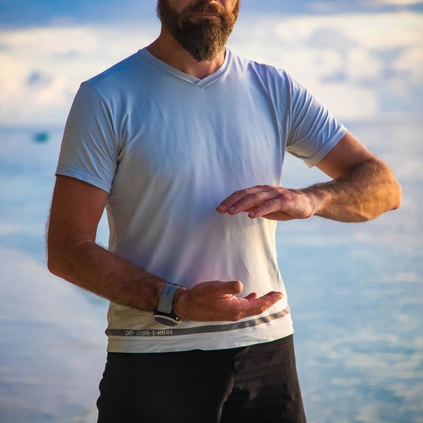 A man with a beard is wearing a white shirt and black shorts, representing a Qi Gong position