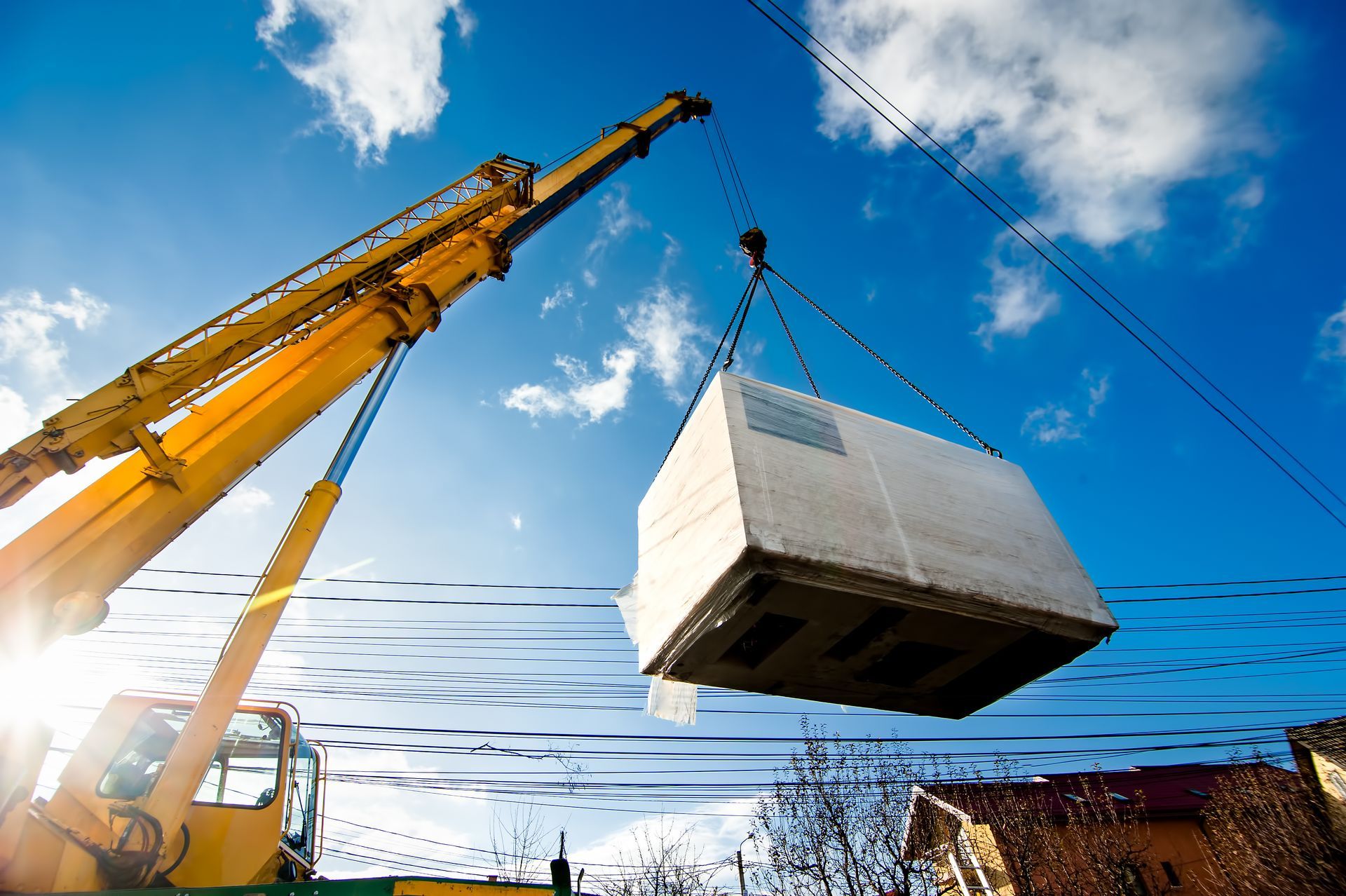 Une grue de chantier jaune soulève un gros bloc de béton rectangulaire sur fond de ciel bleu éclatant et ensoleillé.