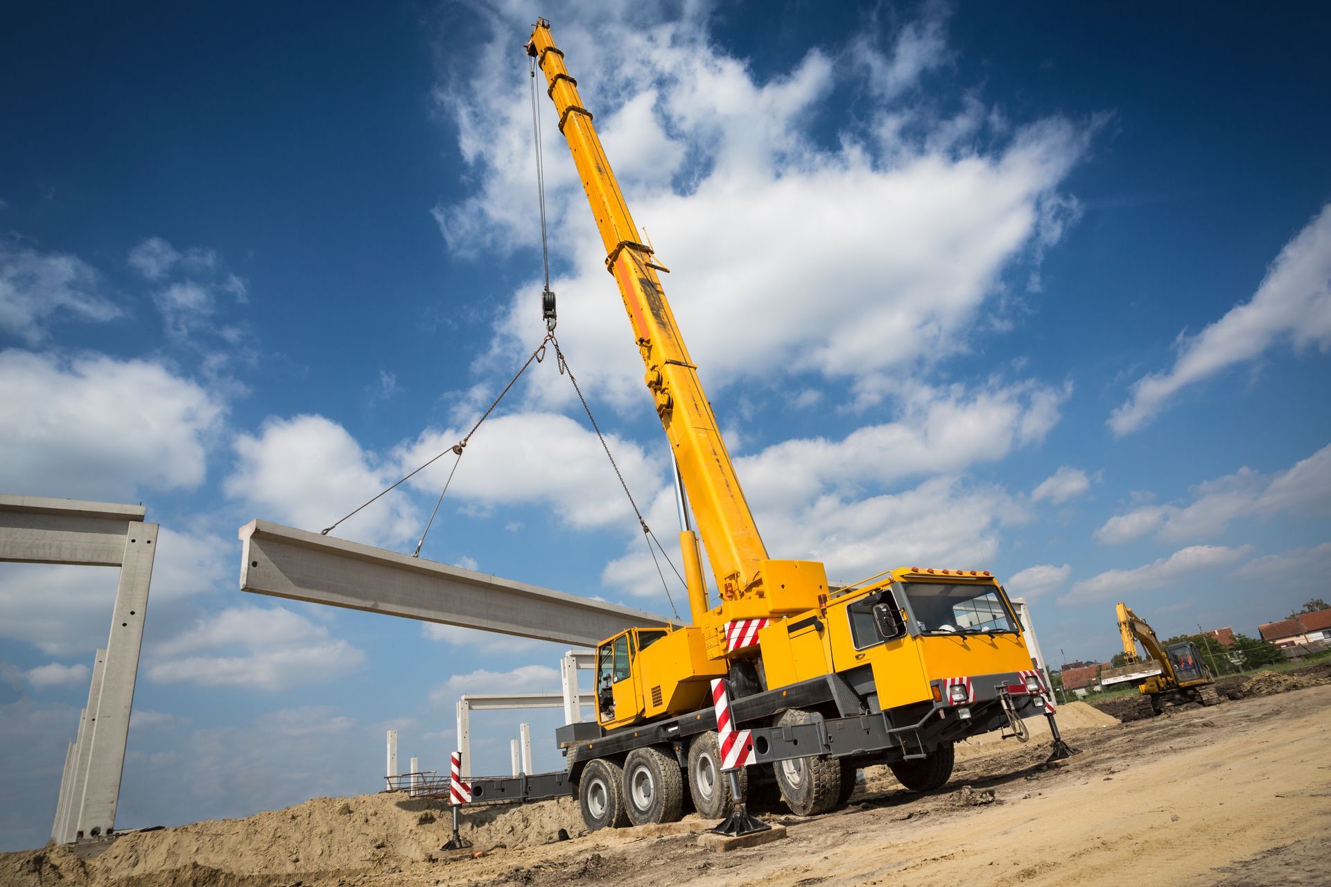 Une grue mobile jaune soulève une grosse poutre en béton sur un chantier de construction sous un ciel bleu nuageux.